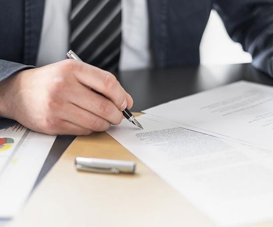 Un hombre vestido de traje firma un documento en un escritorio, con papeles y un bolígrafo cerca.