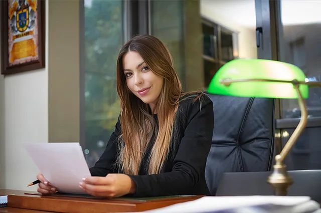 Mujer sentada en un escritorio con papeles en la mano en una oficina, con una lámpara de escritorio verde y un ordenador portátil cerca.