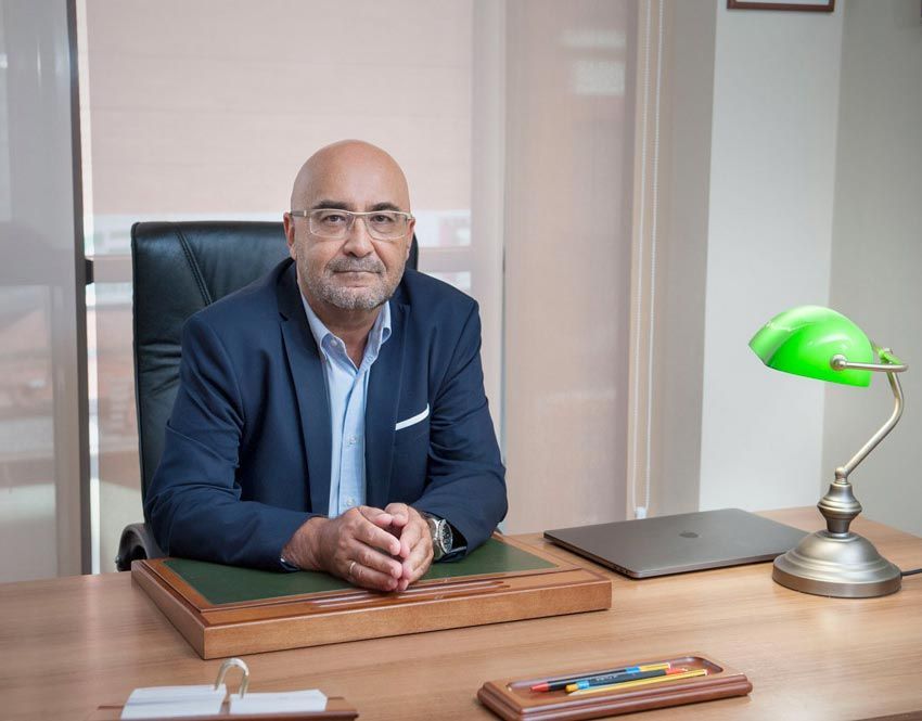 Man seated at a desk in an office, wearing a navy blazer, with a green desk lamp and laptop nearby