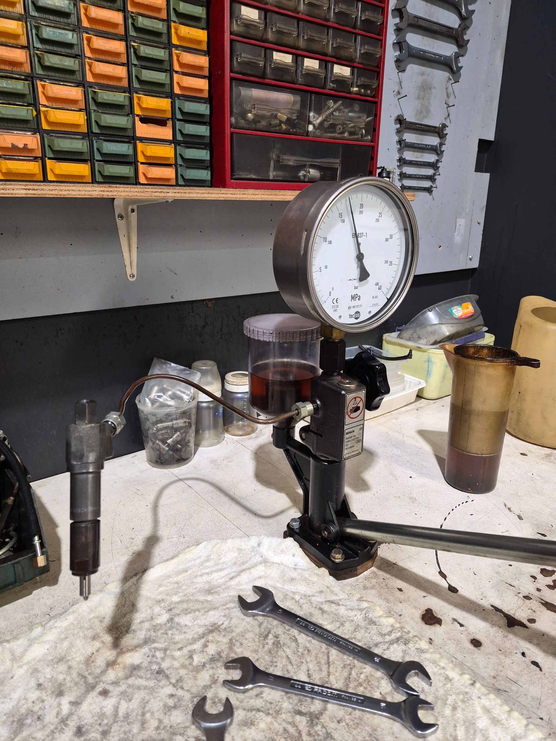 Un testeur d'injecteurs de carburant diesel avec un manomètre, un injecteur et des clés sur un établi.