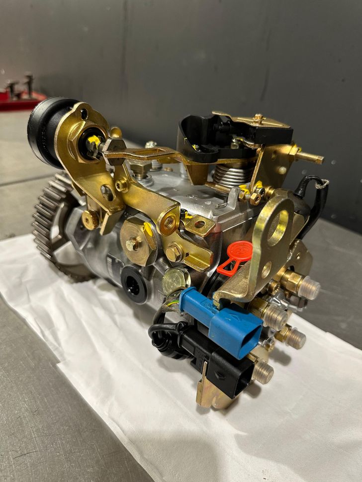 Pompe d'injection de carburant avec divers composants métalliques et connecteurs électriques sur une surface blanche.