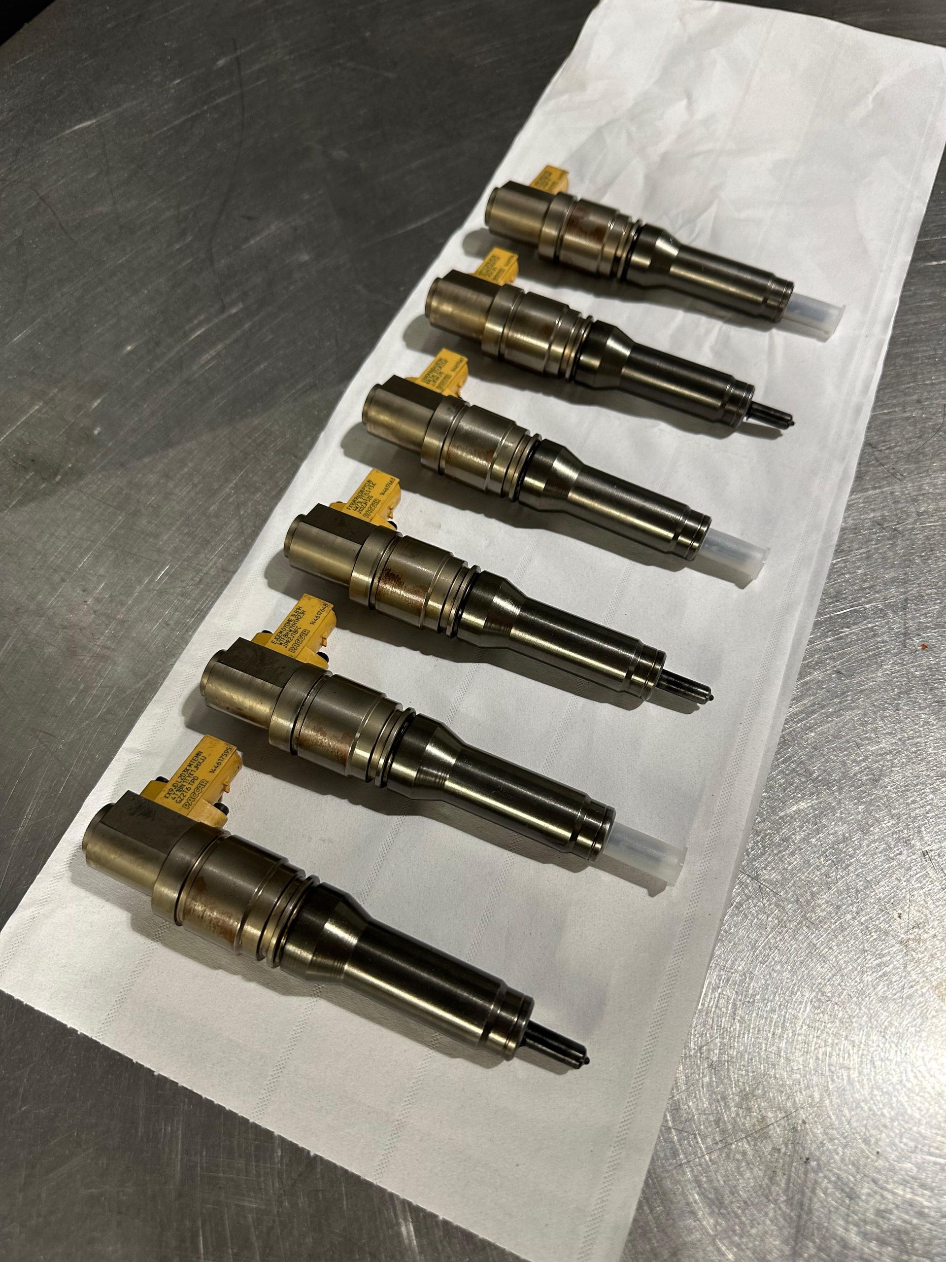 Six injecteurs de carburant, métalliques et noirs, avec des connecteurs jaunes, disposés sur une serviette en papier blanche, sur une surface métallique.
