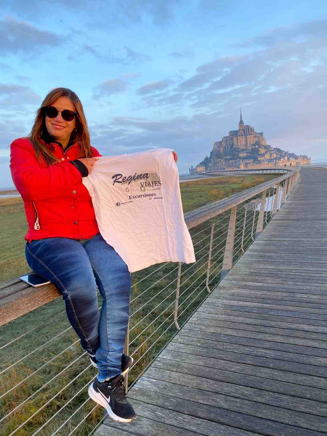 Mujer con chaqueta roja sostiene una camisa blanca, sentada en un paseo marítimo con el Mont Saint-Michel al fondo.