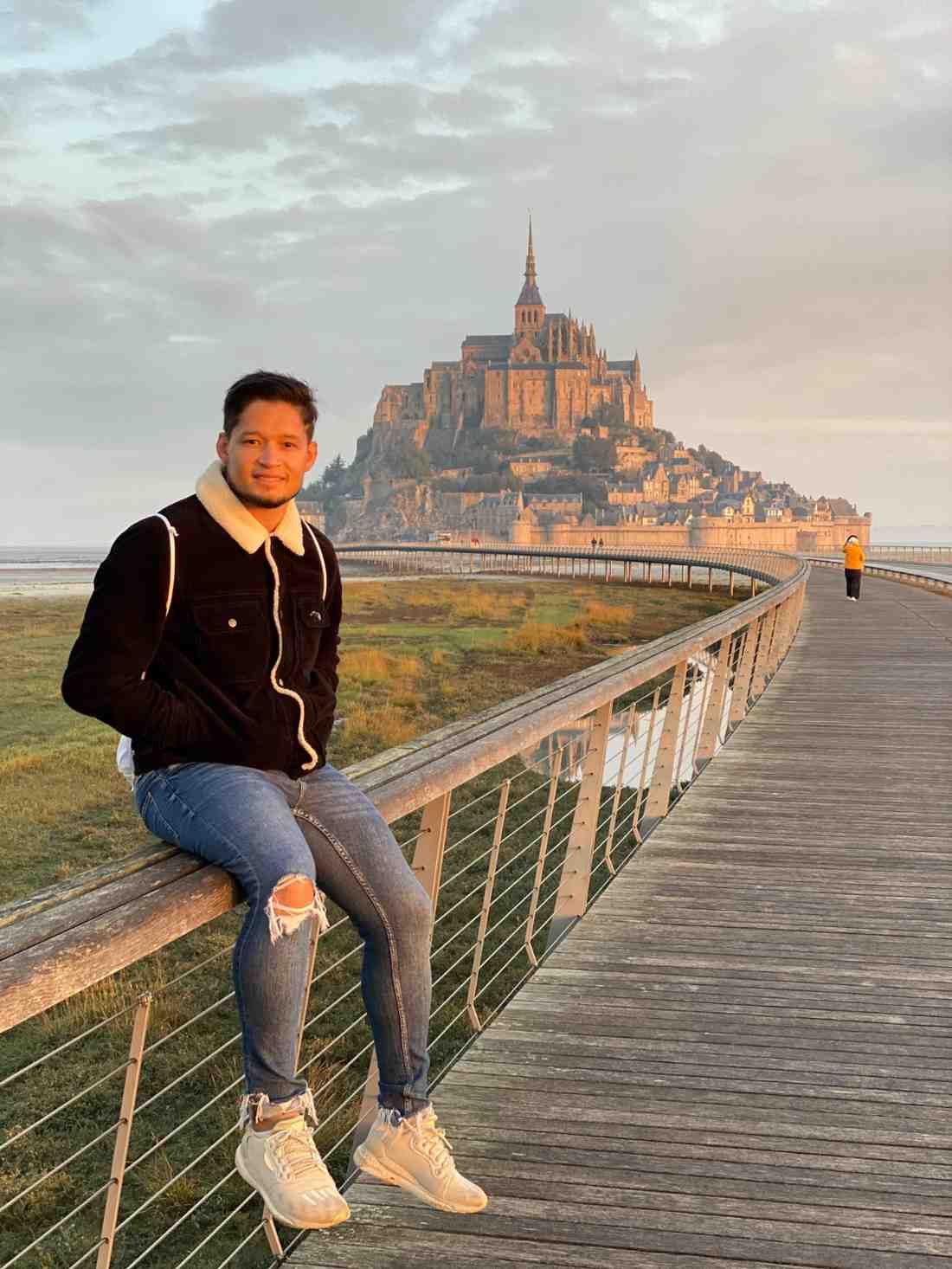Hombre sentado en una barandilla de madera, con el Mont Saint-Michel al fondo, vaqueros azules, chaqueta negra, día soleado.