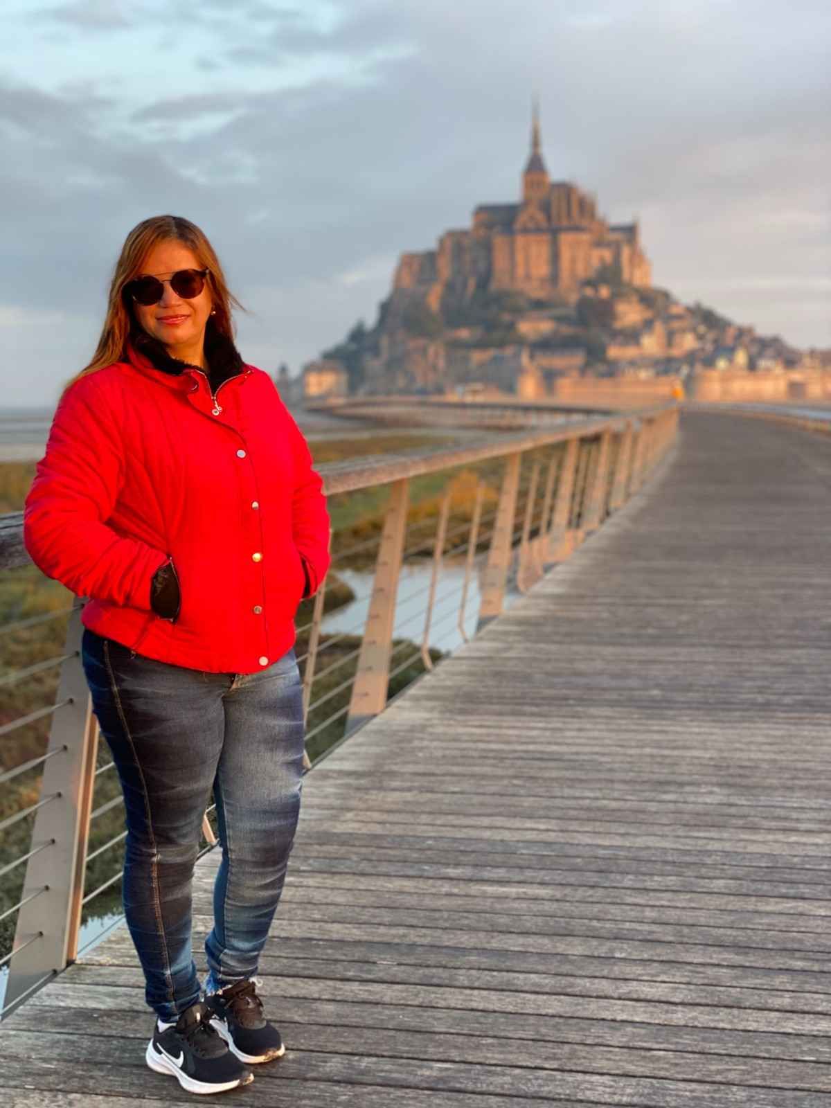 Una mujer con chaqueta roja posa en un puente con el Mont Saint-Michel al fondo.