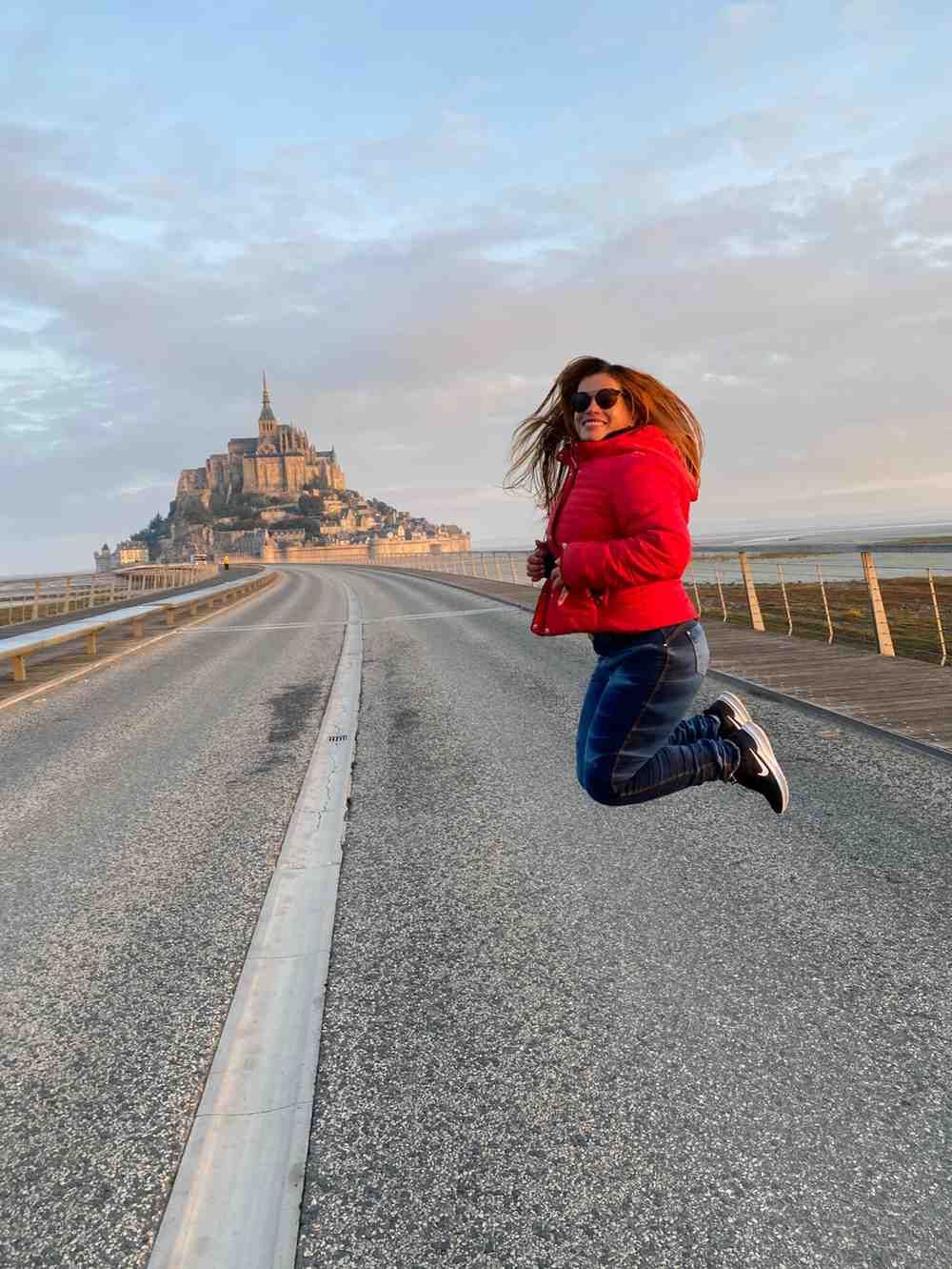 Mujer saltando en una carretera con el Mont Saint-Michel al fondo, vistiendo una chaqueta roja y jeans azules.