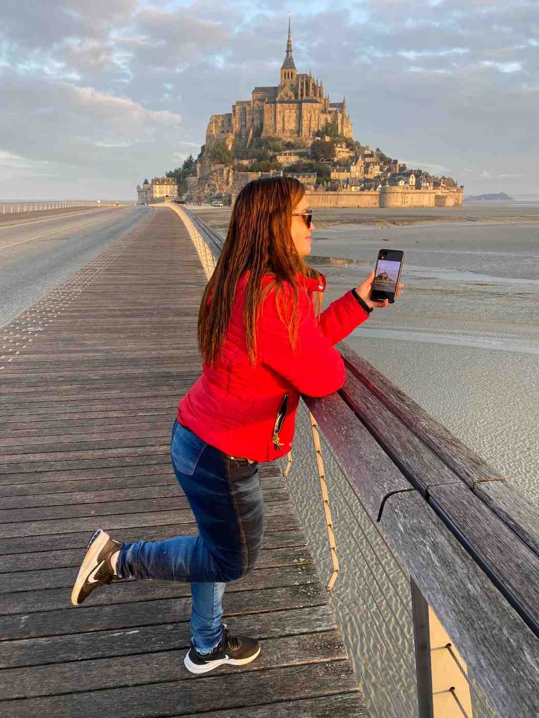 Mujer tomándose un selfie con el Mont Saint-Michel al fondo, de pie sobre una pasarela de madera.