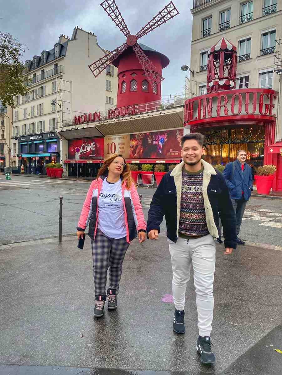 Una pareja de la mano camina frente al Moulin Rouge en París. Edificios y cielo nublado al fondo.