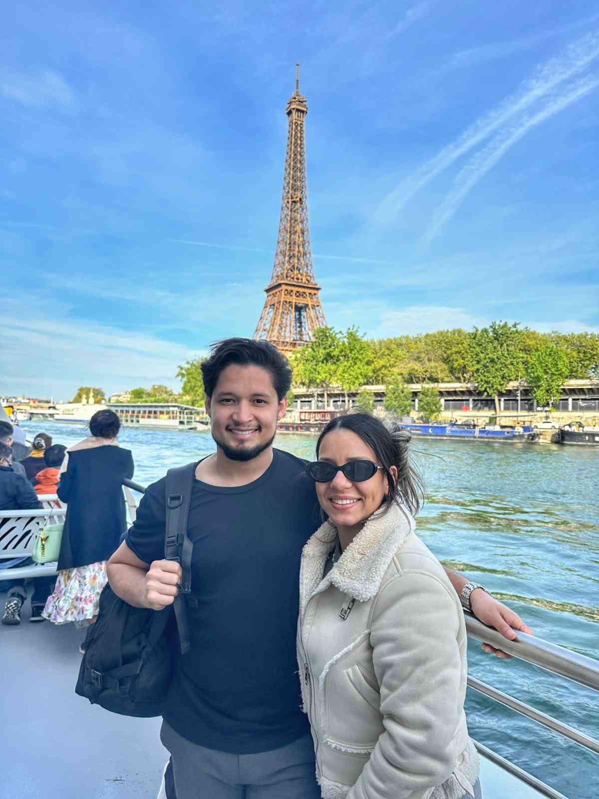 Pareja en un barco con la Torre Eiffel al fondo, cielo azul.