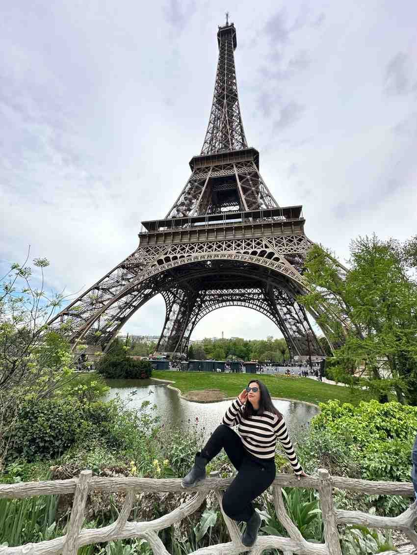 Mujer posando en una valla frente a la Torre Eiffel, vestida con pantalones negros y un suéter de rayas.