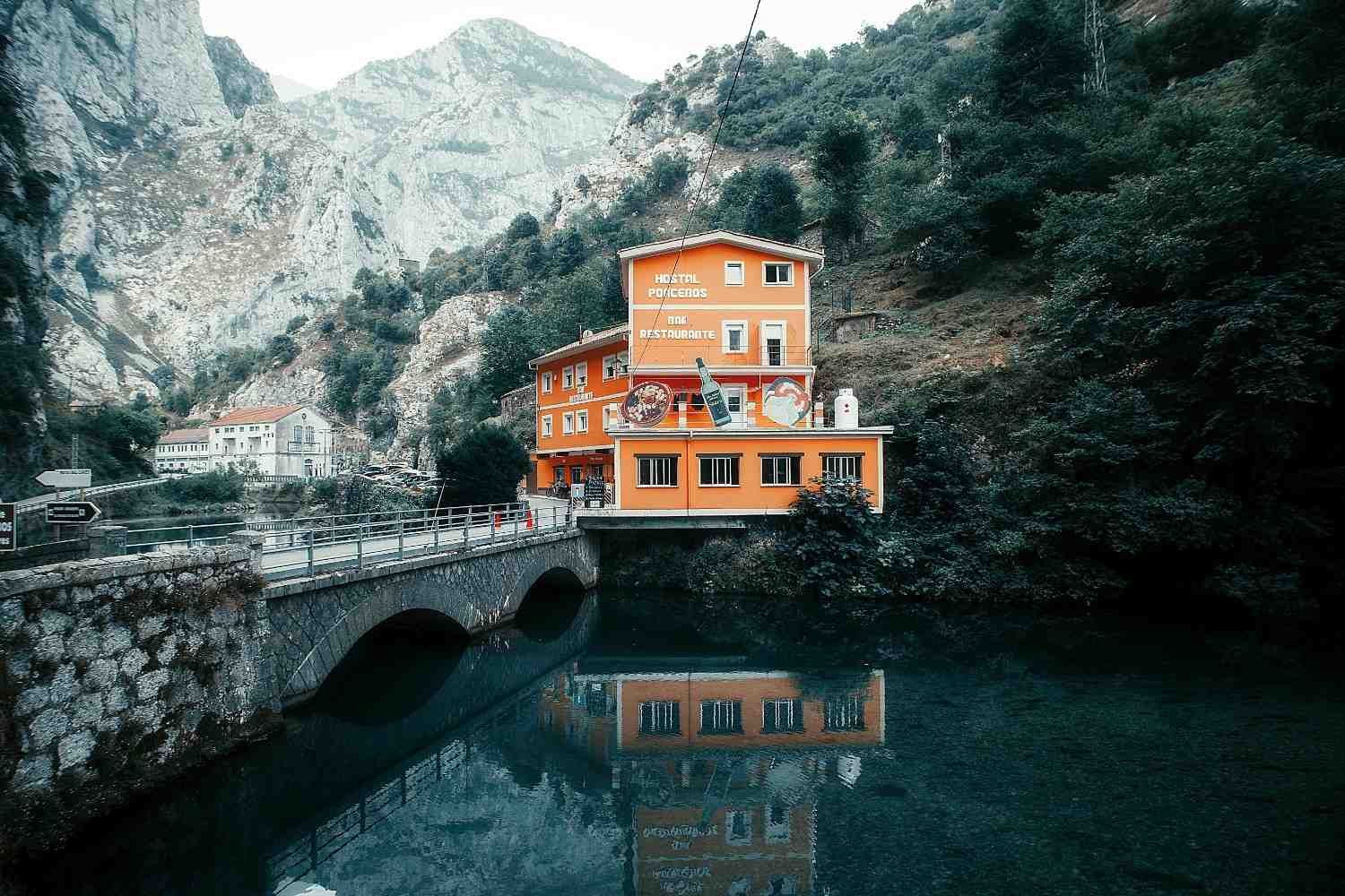 Edificio naranja en la orilla del río, reflejado en el agua, con puente y montaña de fondo.