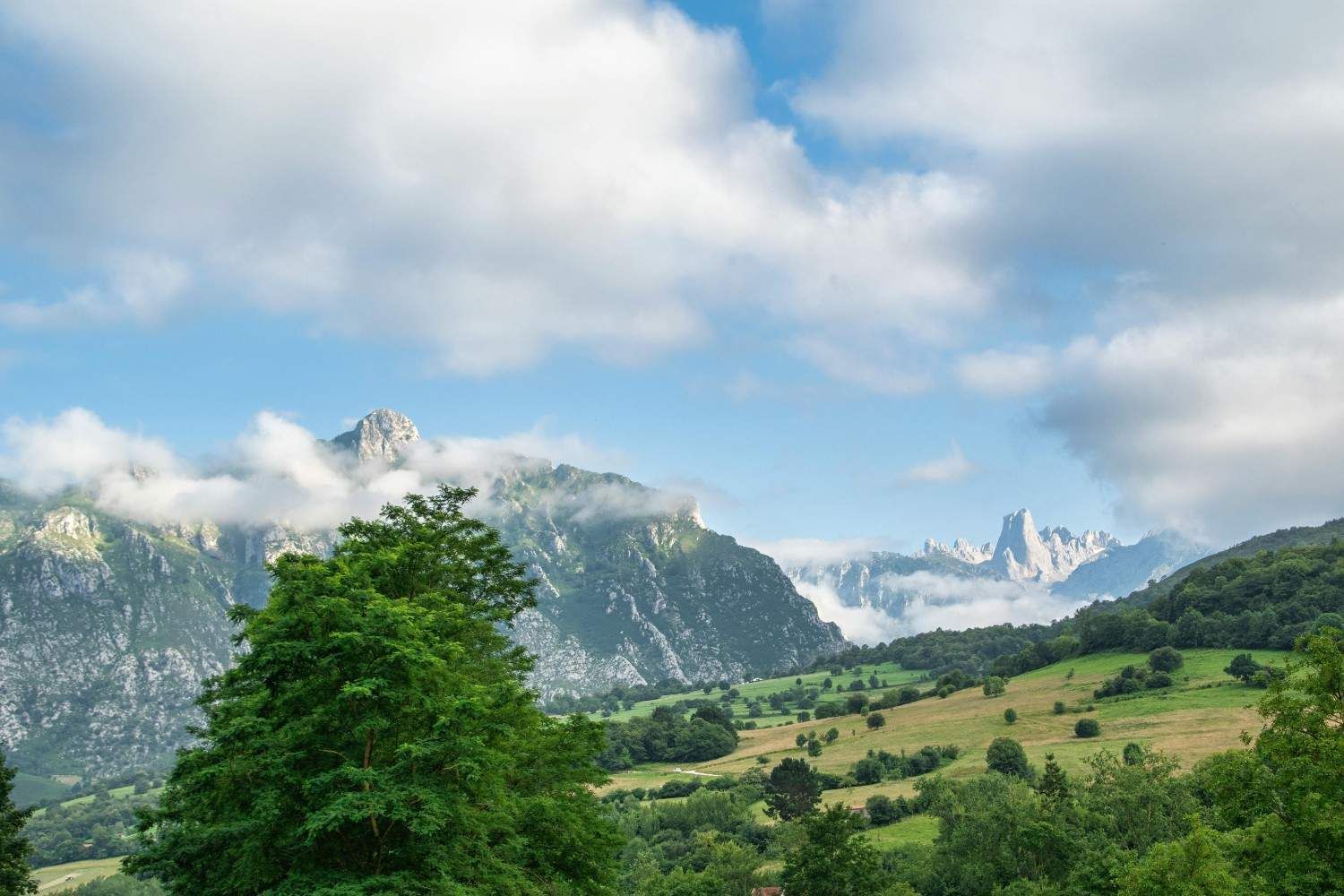 Las montañas se elevan sobre un valle verde, parcialmente oscurecido por las nubes, bajo un cielo azul.