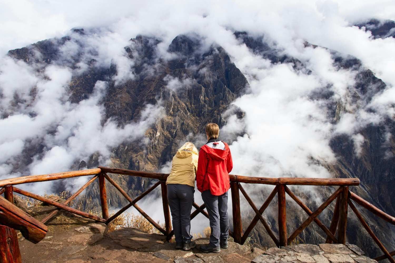 Pareja admirando una vista de montaña llena de nubes desde un mirador de madera, uno de ellos vistiendo una chaqueta roja.