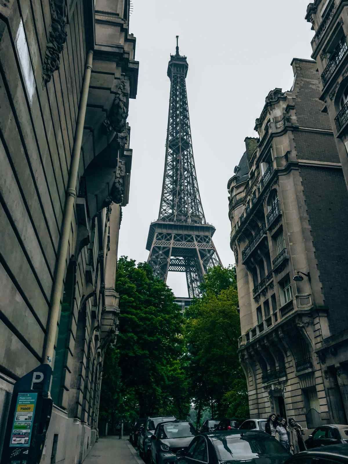 La Torre Eiffel enmarcada por edificios, París. Coches en la calle, cielo nublado, árboles verdes.