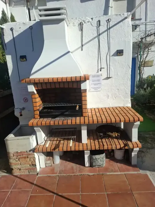 Barbacoa de ladrillo con chimenea frente a un edificio blanco. Estante para utensilios de parrilla. Patio de baldosas rojas.