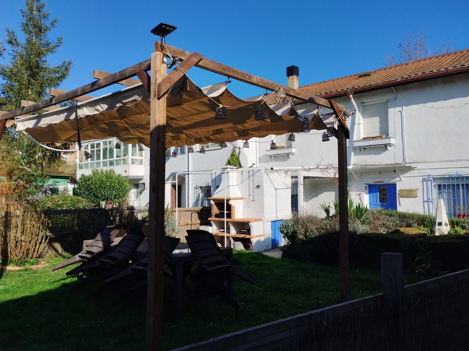 Pérgola de madera con sombrilla en un patio, con vista a un edificio blanco con una puerta azul.