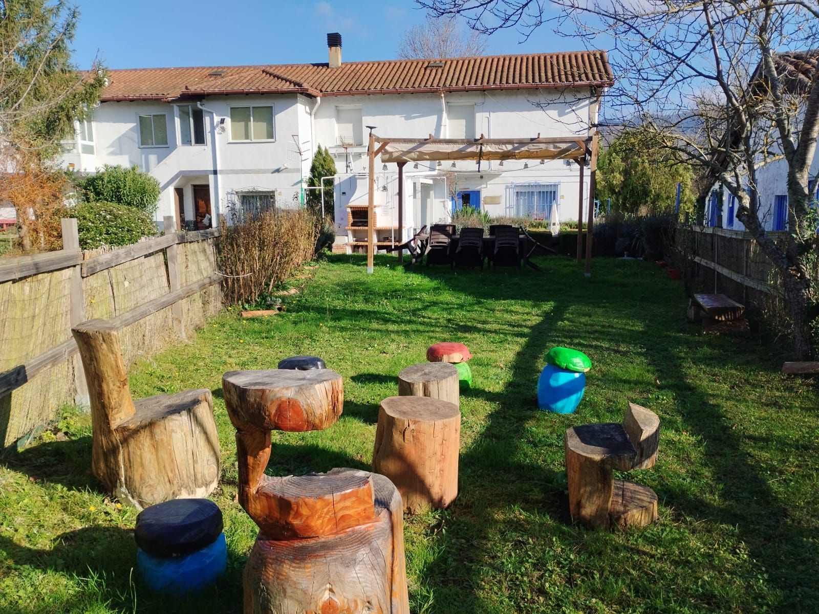 Patio trasero con bancos de troncos de árboles, césped, cerca de madera y una casa blanca. Pérgola con sillas al fondo.