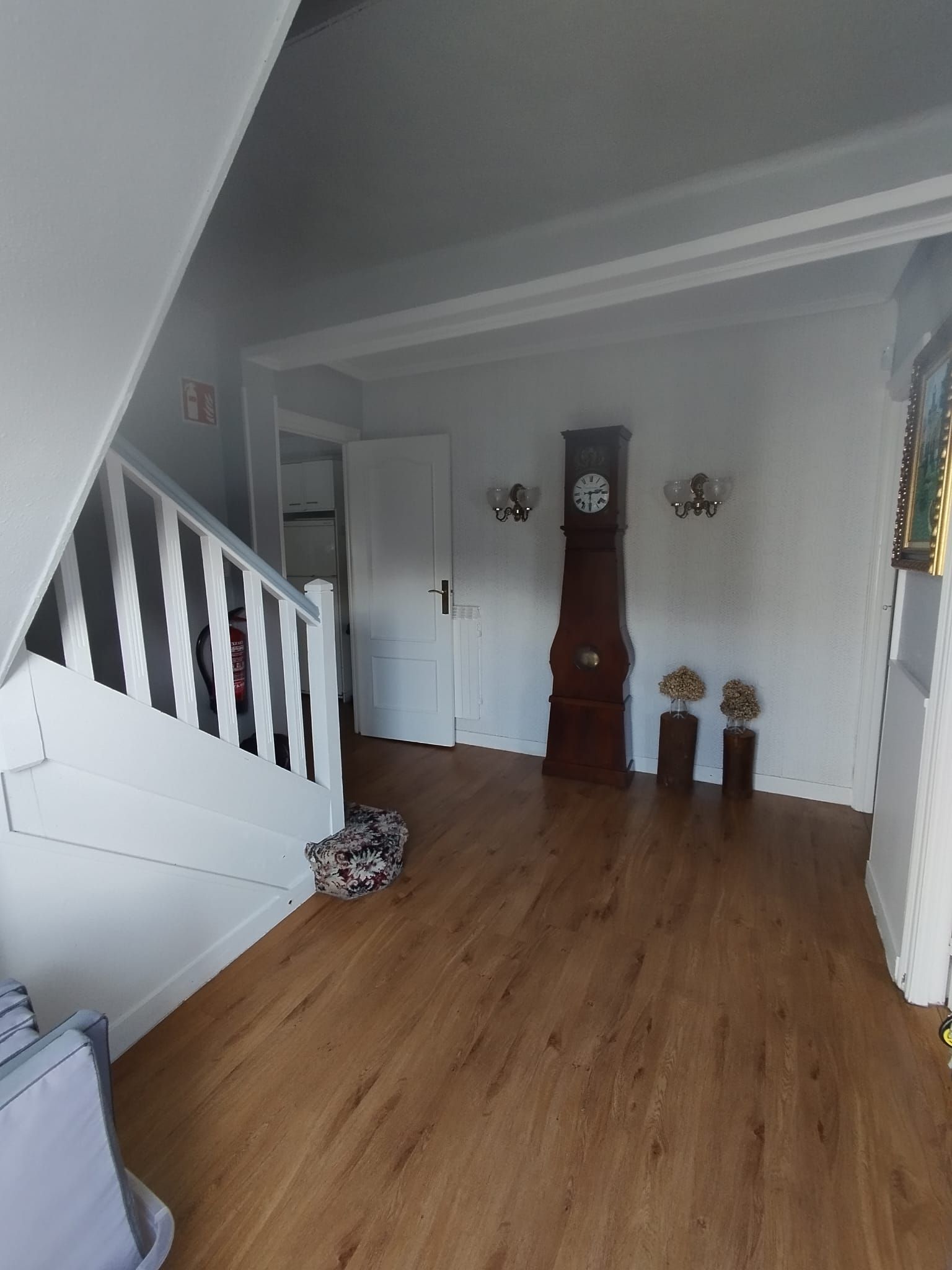 Pasillo con suelo de madera, escalera a la izquierda, reloj de pie y dos apliques. Paredes y puerta blancas.