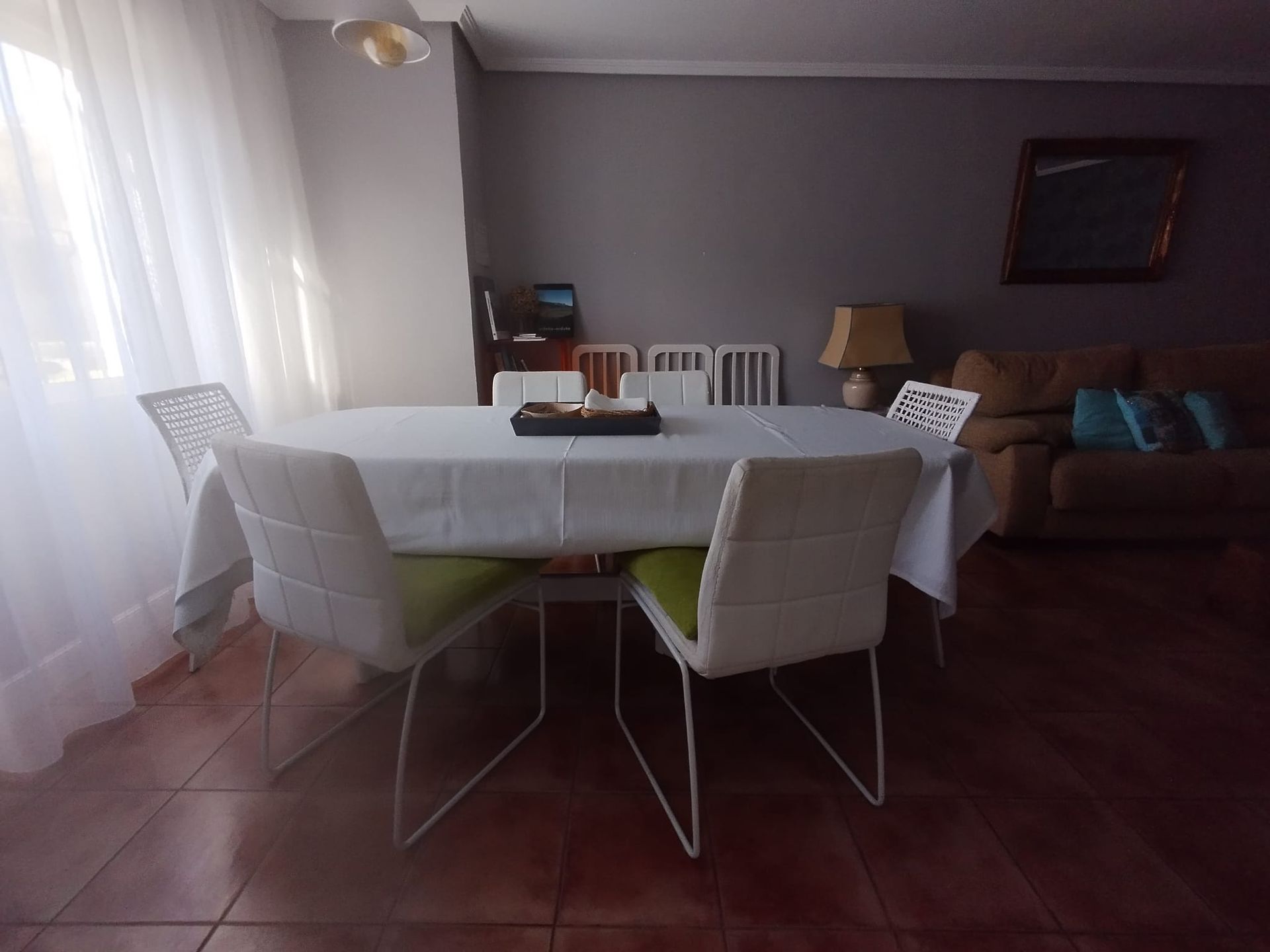Comedor: mesa con sillas blancas, cubierta con un mantel blanco. Suelo de baldosas marrones, paredes grises, luz solar.