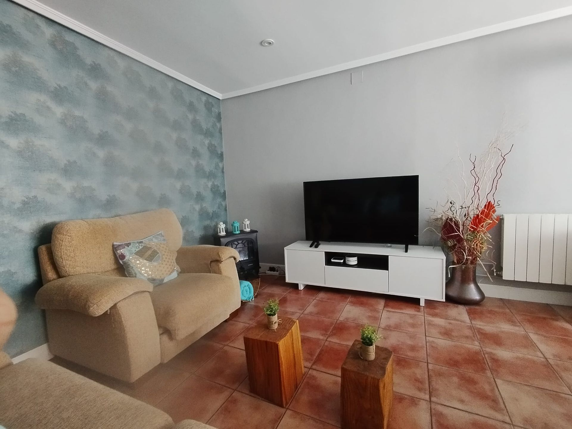 Sala de estar con sofá beige, televisor y mesas de centro de madera sobre suelo de baldosas. Pared con acento azul.