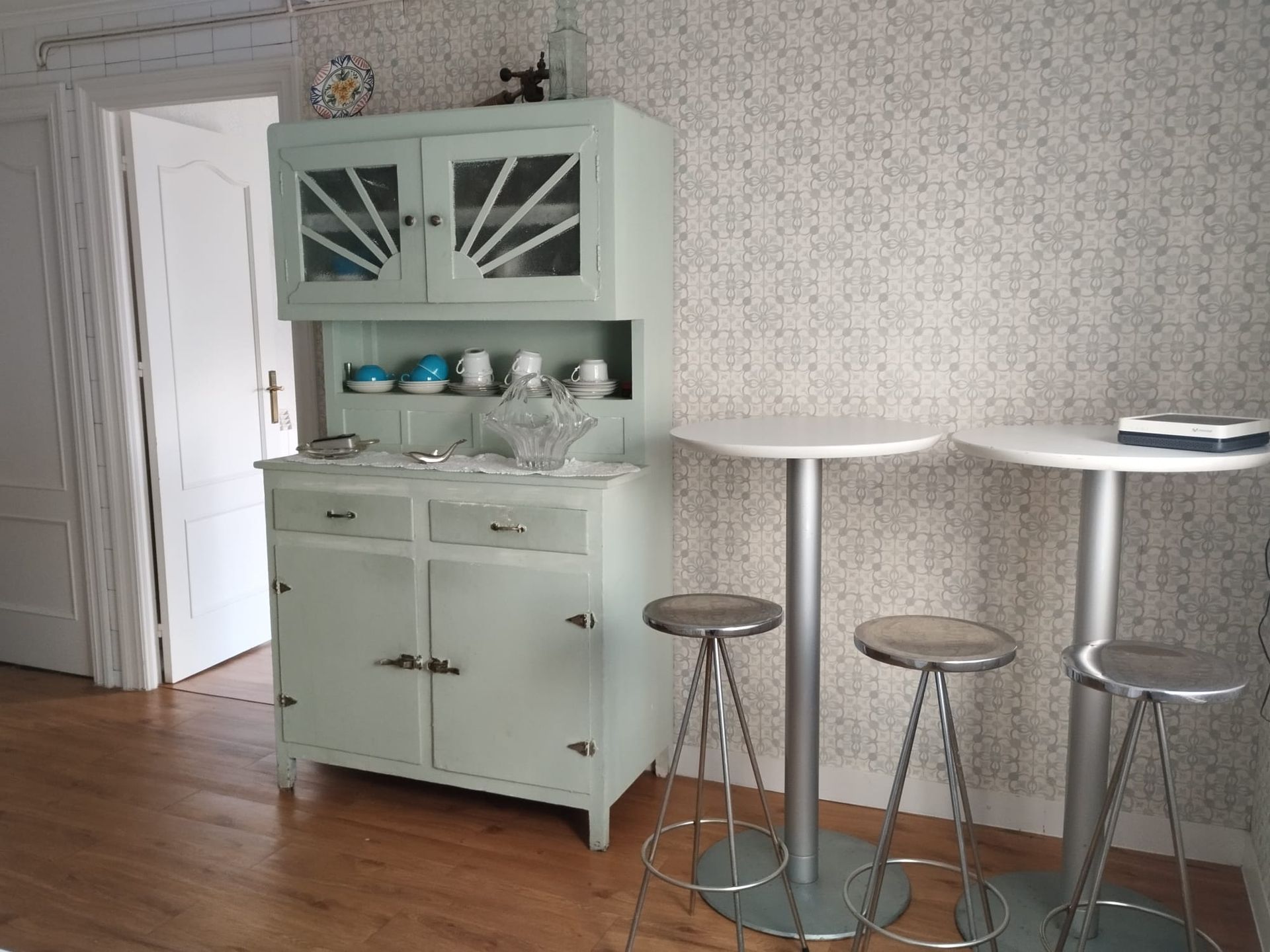 Un aparador verde claro con platos, dos mesas de bistró blancas y taburetes en una habitación con papel tapiz estampado.