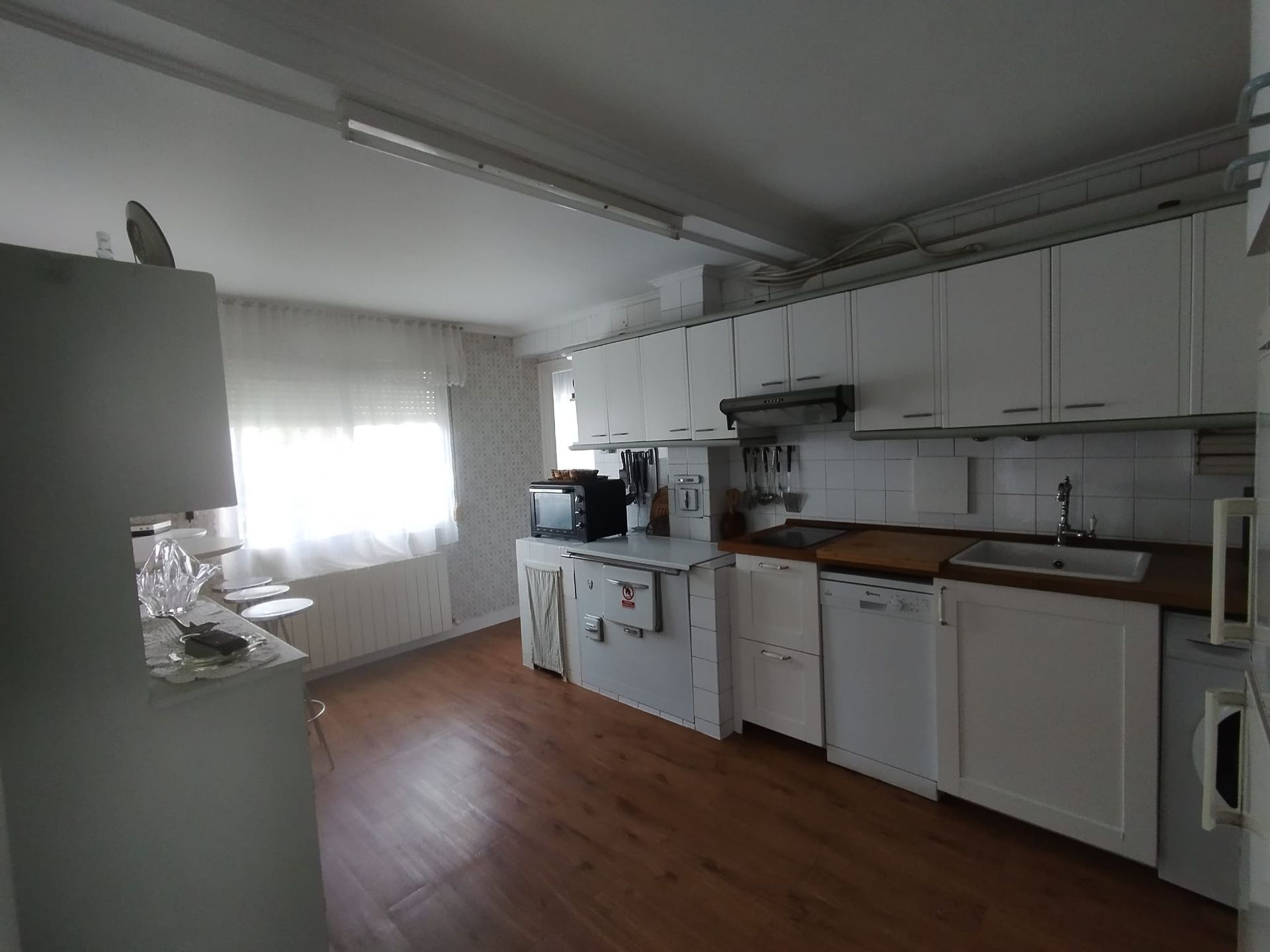 Cocina blanca con armarios, electrodomésticos y ventana. Suelo de madera.