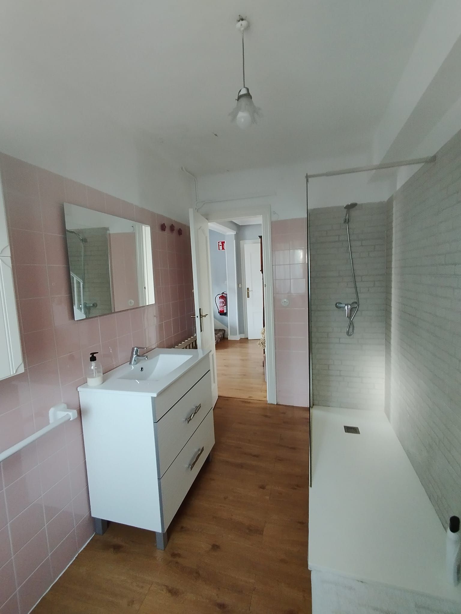 Baño con tocador blanco, espejo, ducha y paredes de azulejos rosa claro. Suelo de madera.