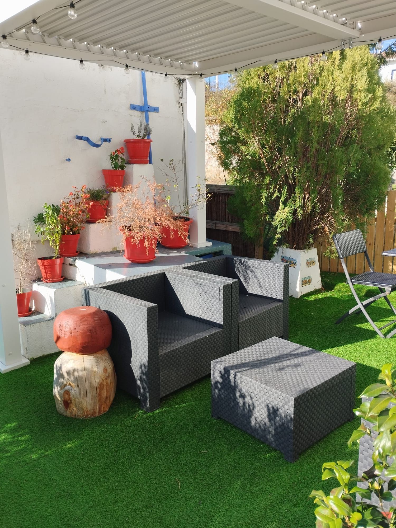 Zona de estar al aire libre con muebles de mimbre negro, plantas en macetas rojas y césped verde.