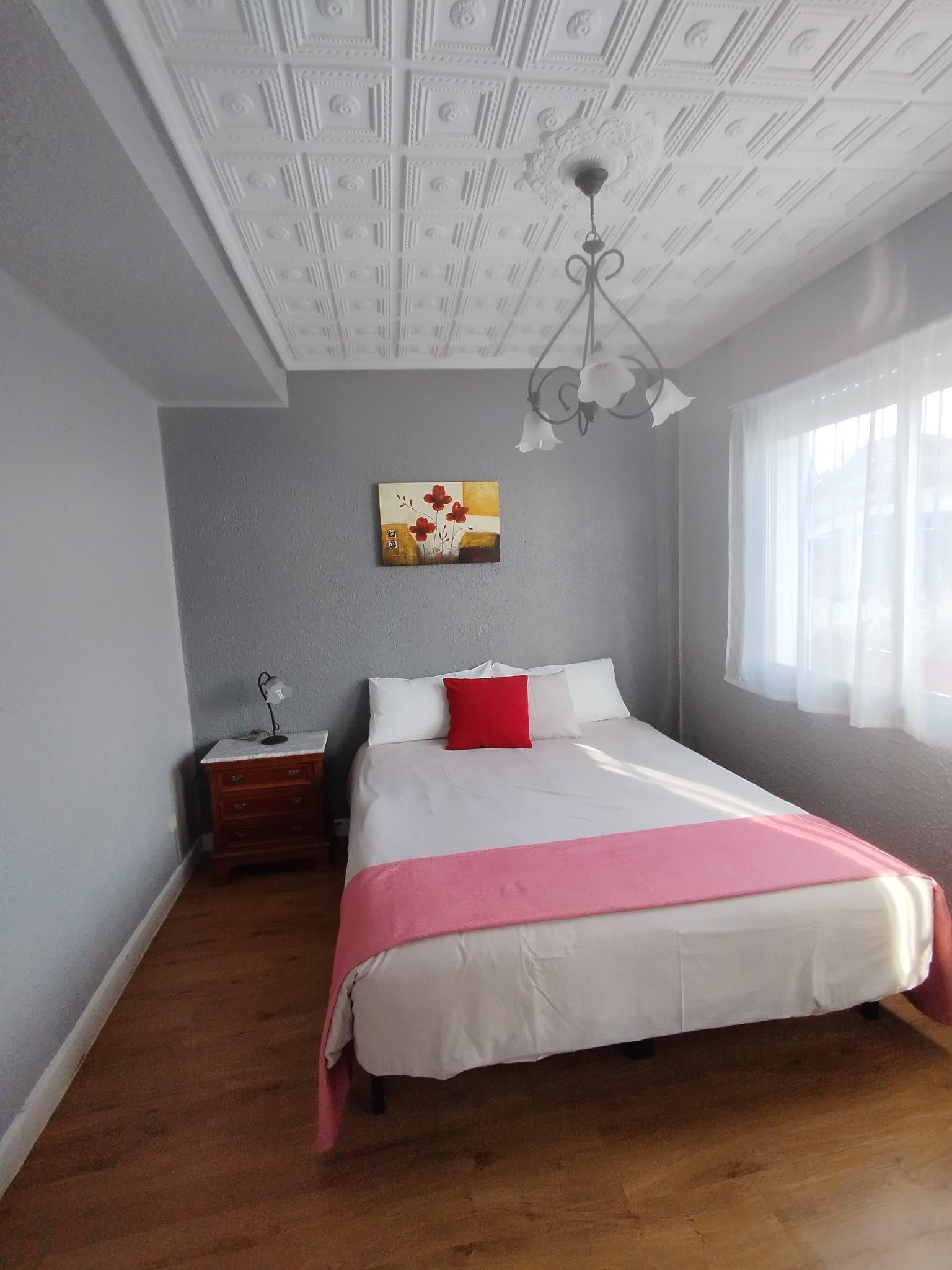 Dormitorio con cama, mesita de noche, obras de arte y lámpara de araña. Paredes grises, suelo de madera y techo blanco.