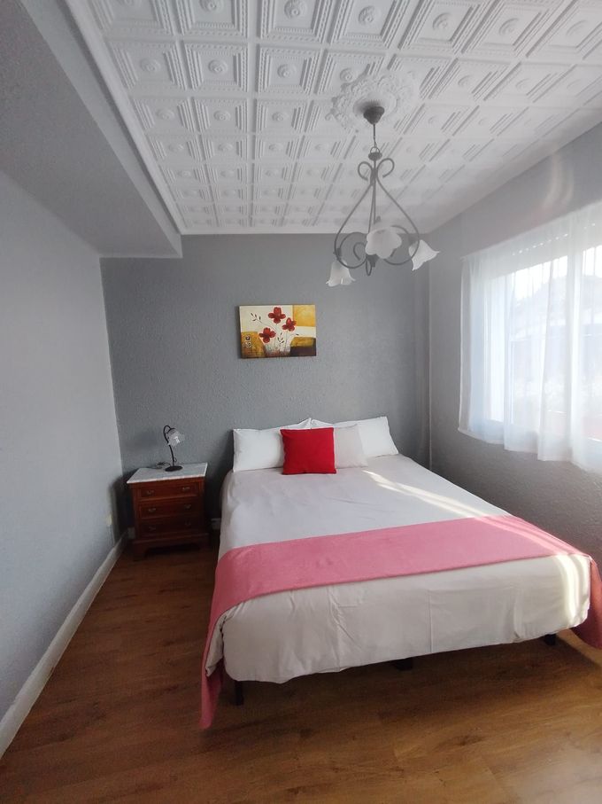 Dormitorio con cama, mesita de noche, obras de arte y lámpara de araña. Paredes grises, suelo de madera y techo blanco.