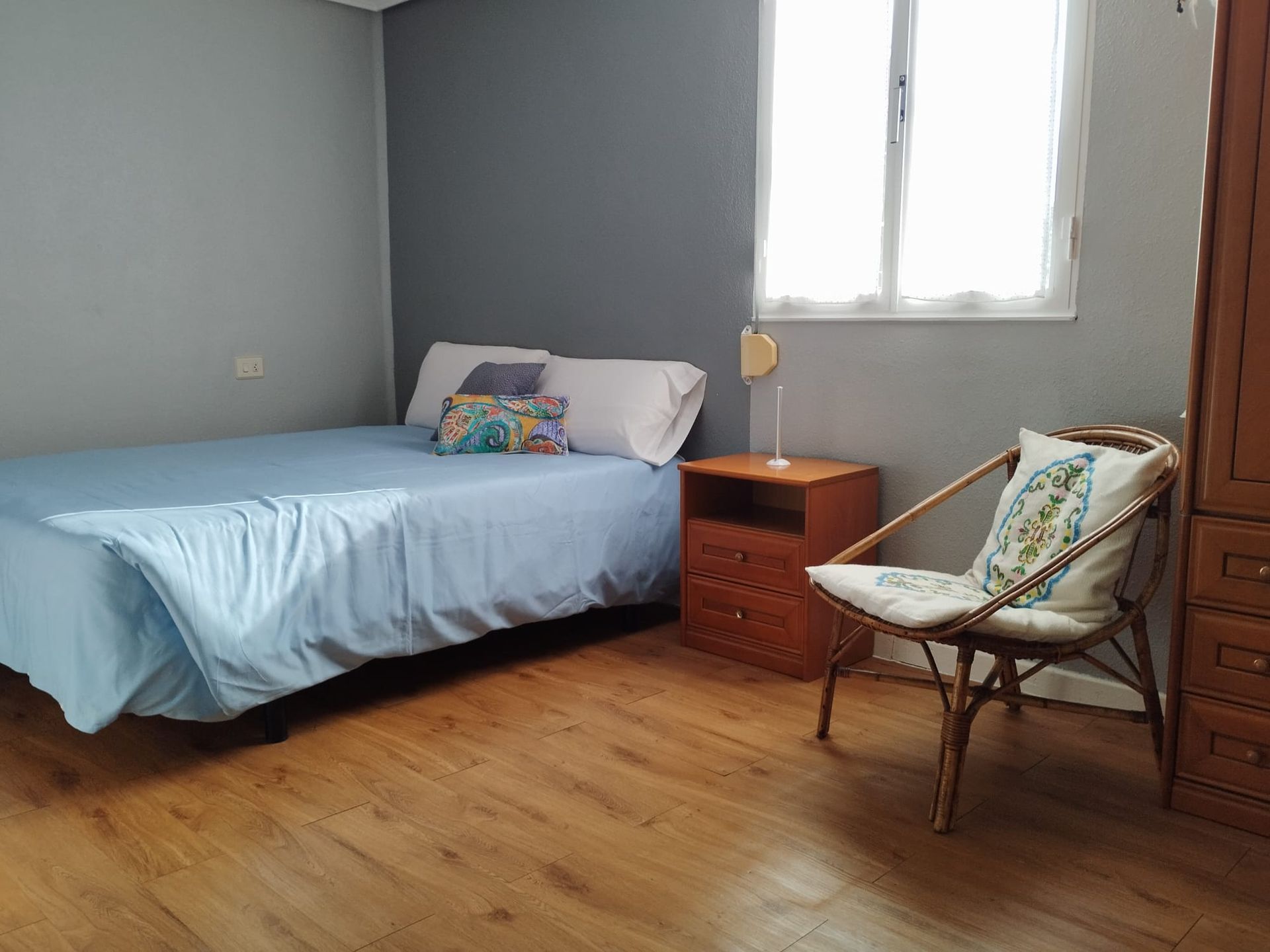 Dormitorio con cama, mesita de noche, silla y armario. Ropa de cama azul, suelo de madera y ventana.