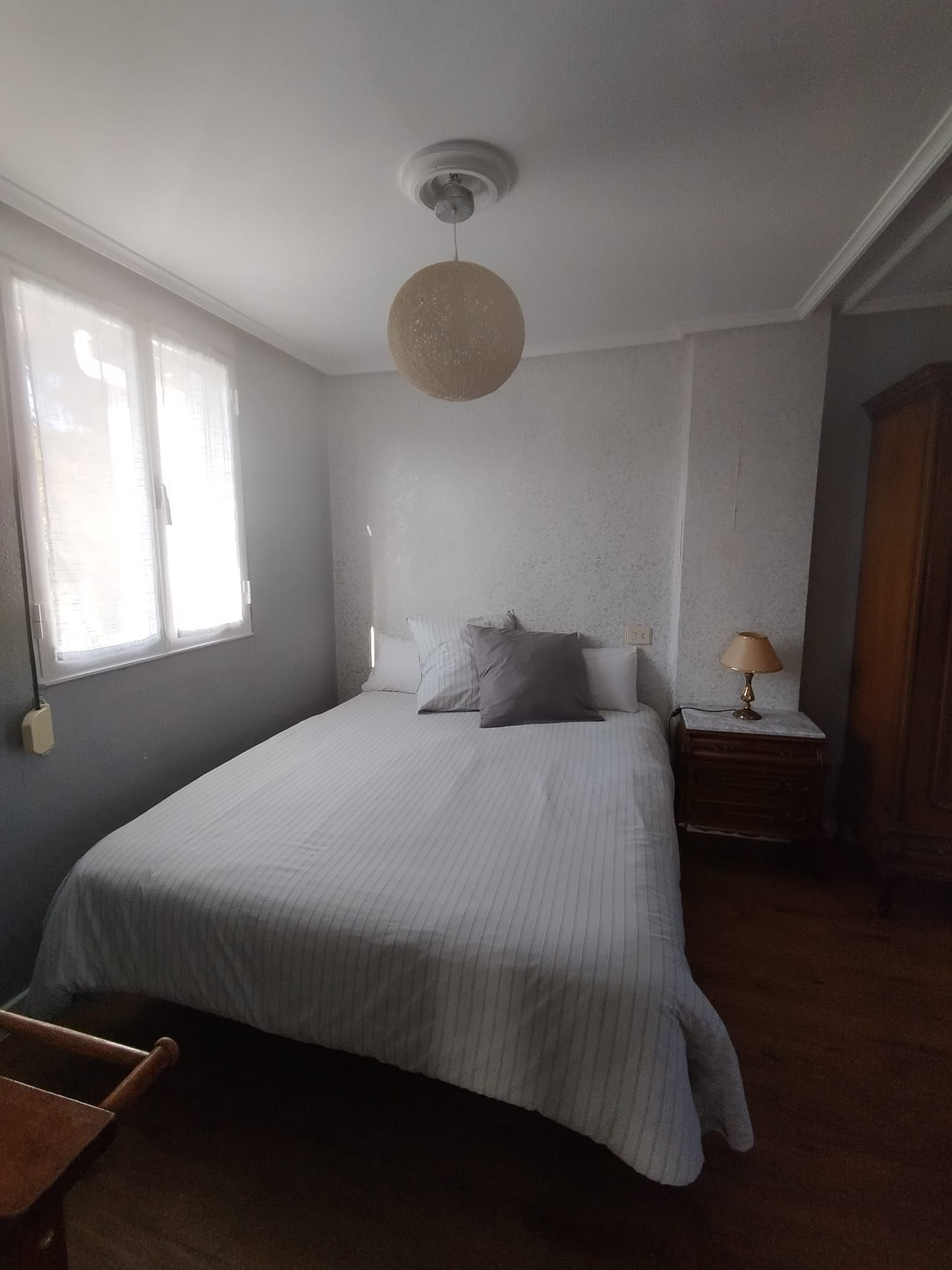 Dormitorio con cama, mesita de noche, armario y lámpara redonda. Una ventana deja entrar la luz del sol.