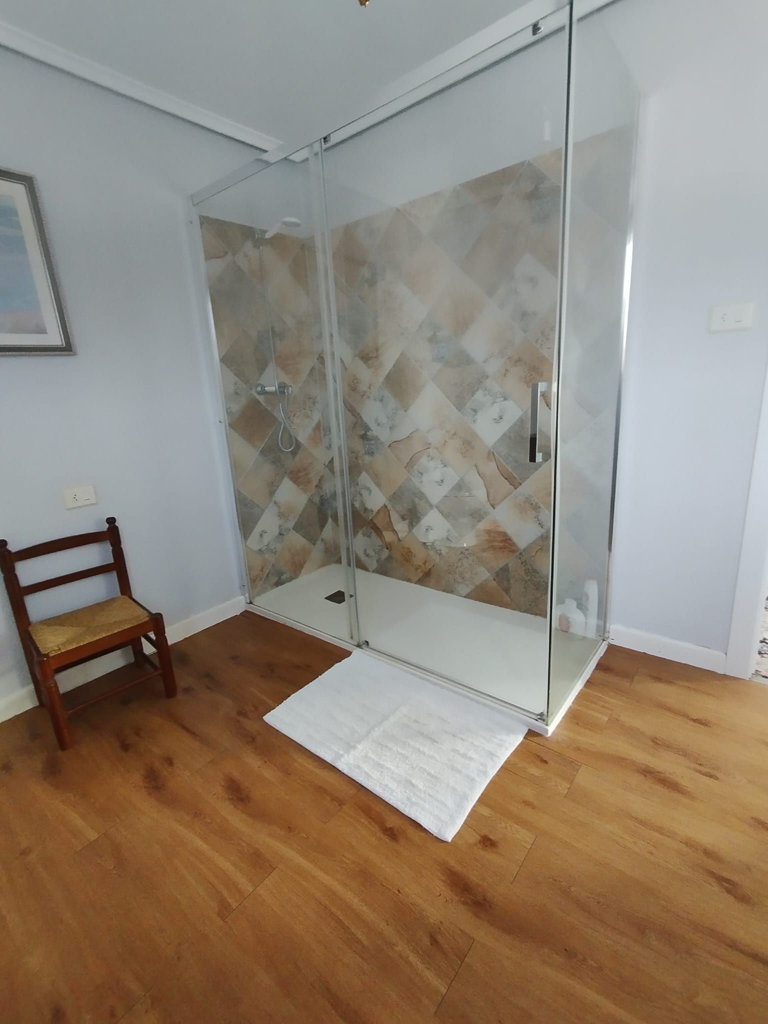 Cabina de ducha con pared posterior de azulejos, puertas de cristal y suelo blanco, junto a una silla de madera.