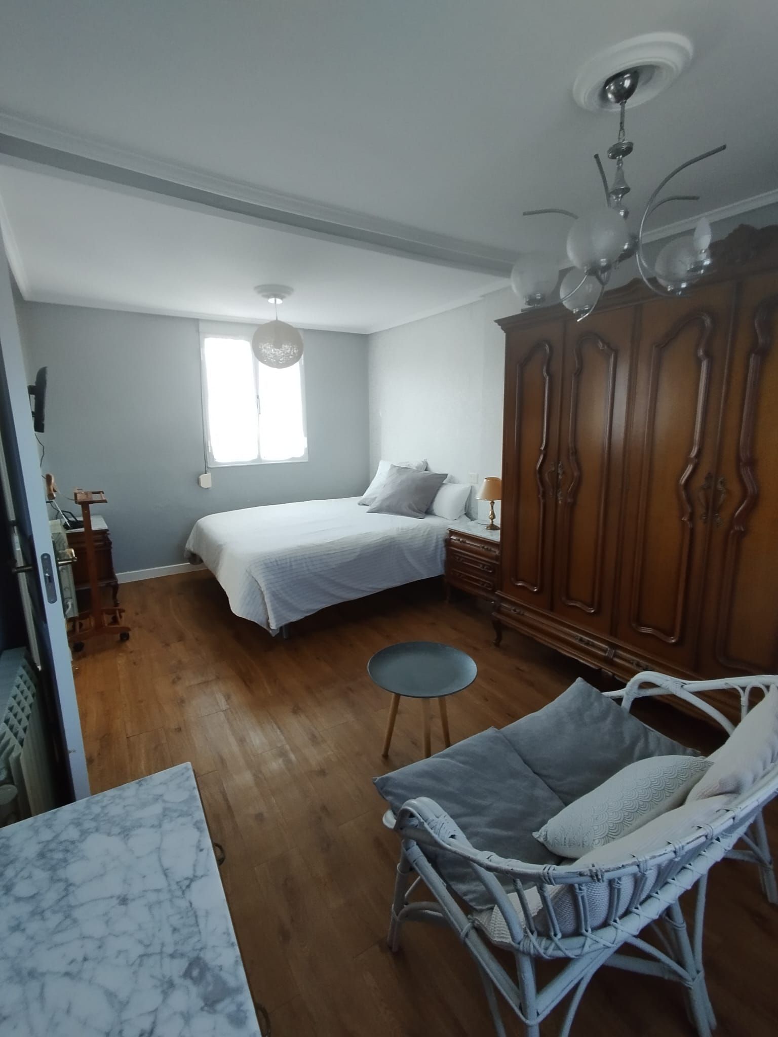 Dormitorio con cama, armario, silla de mimbre y suelo de madera. Paredes de color gris claro.