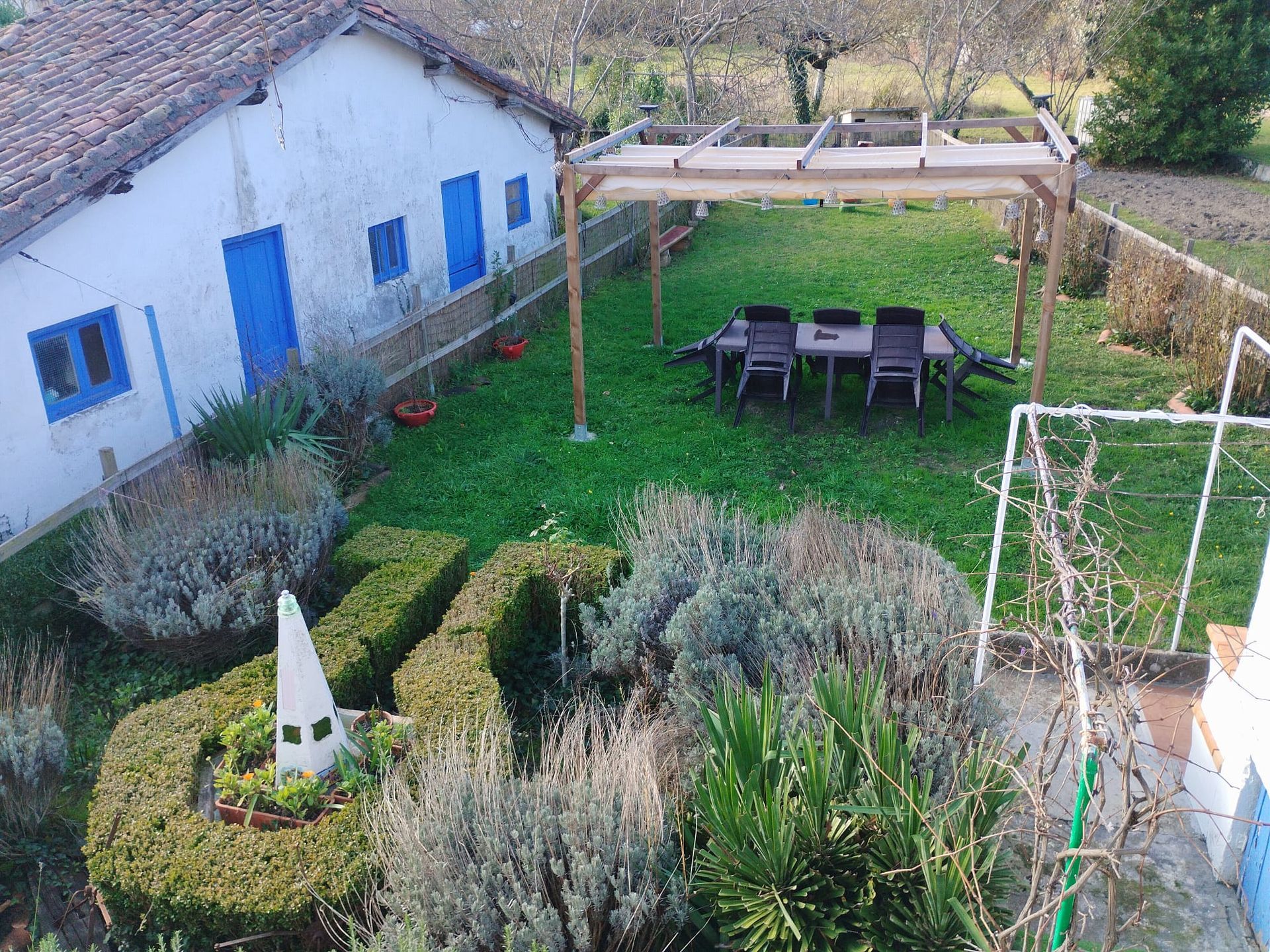 Un patio trasero con una casa blanca, contraventanas azules, una pérgola sobre una mesa, un jardín esculpido y césped verde.