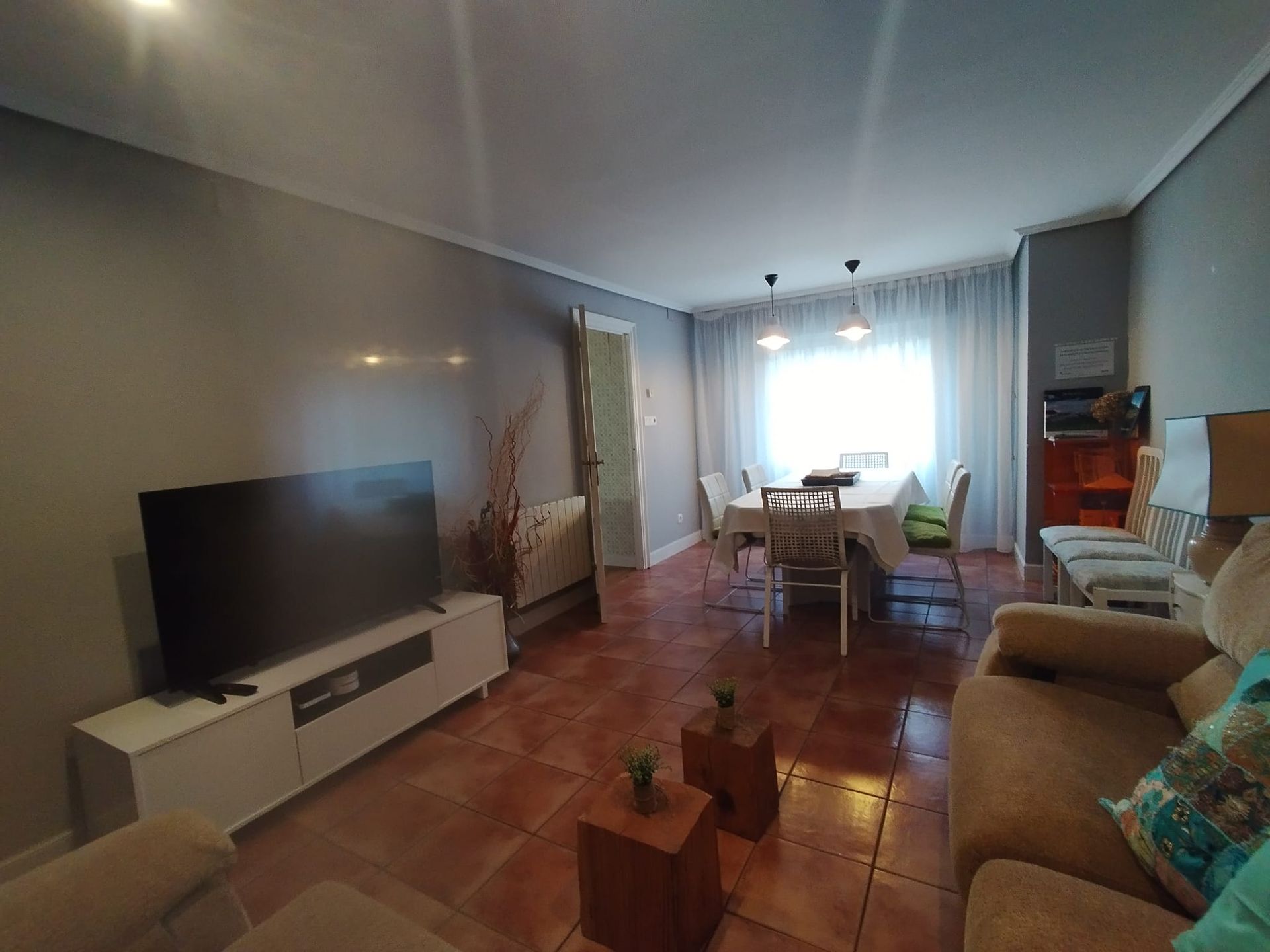 Sala de estar con TV, sofá, mesa de comedor y suelo de baldosas marrones. Paredes grises y molduras blancas.