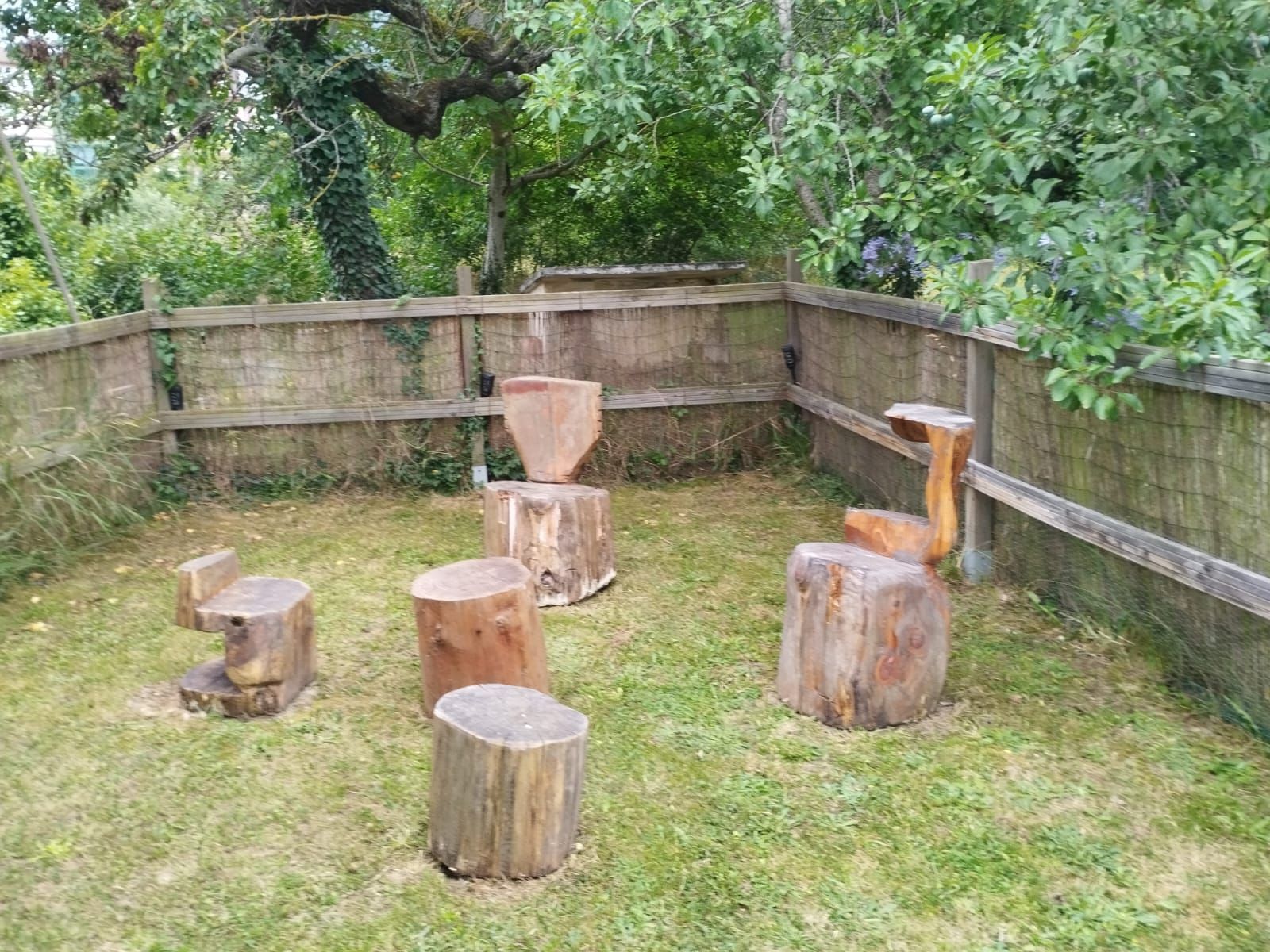 Una zona verde al aire libre con valla de madera y taburetes y sillas de madera tallada.