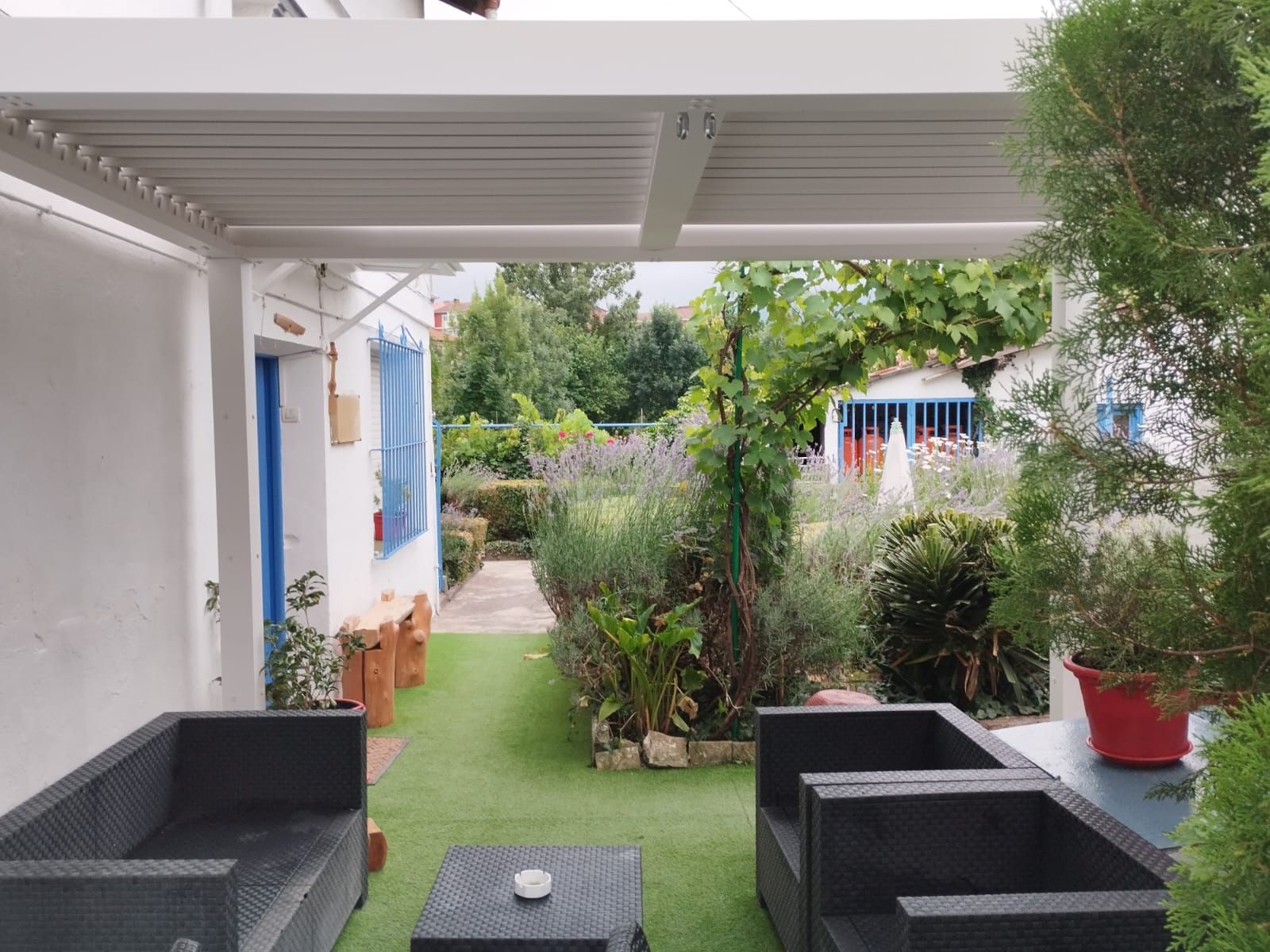 Patio exterior con mobiliario gris, césped artificial y pérgola blanca, que da a un jardín.