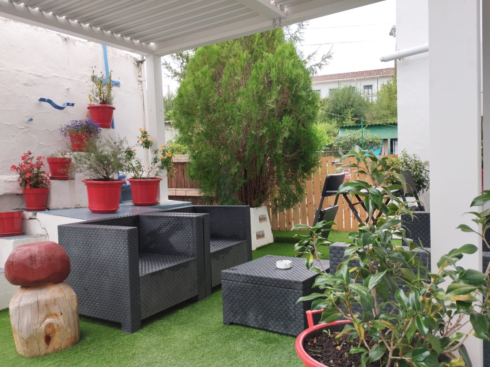 Patio con muebles de mimbre oscuro, plantas en macetas en una pared blanca y césped artificial.