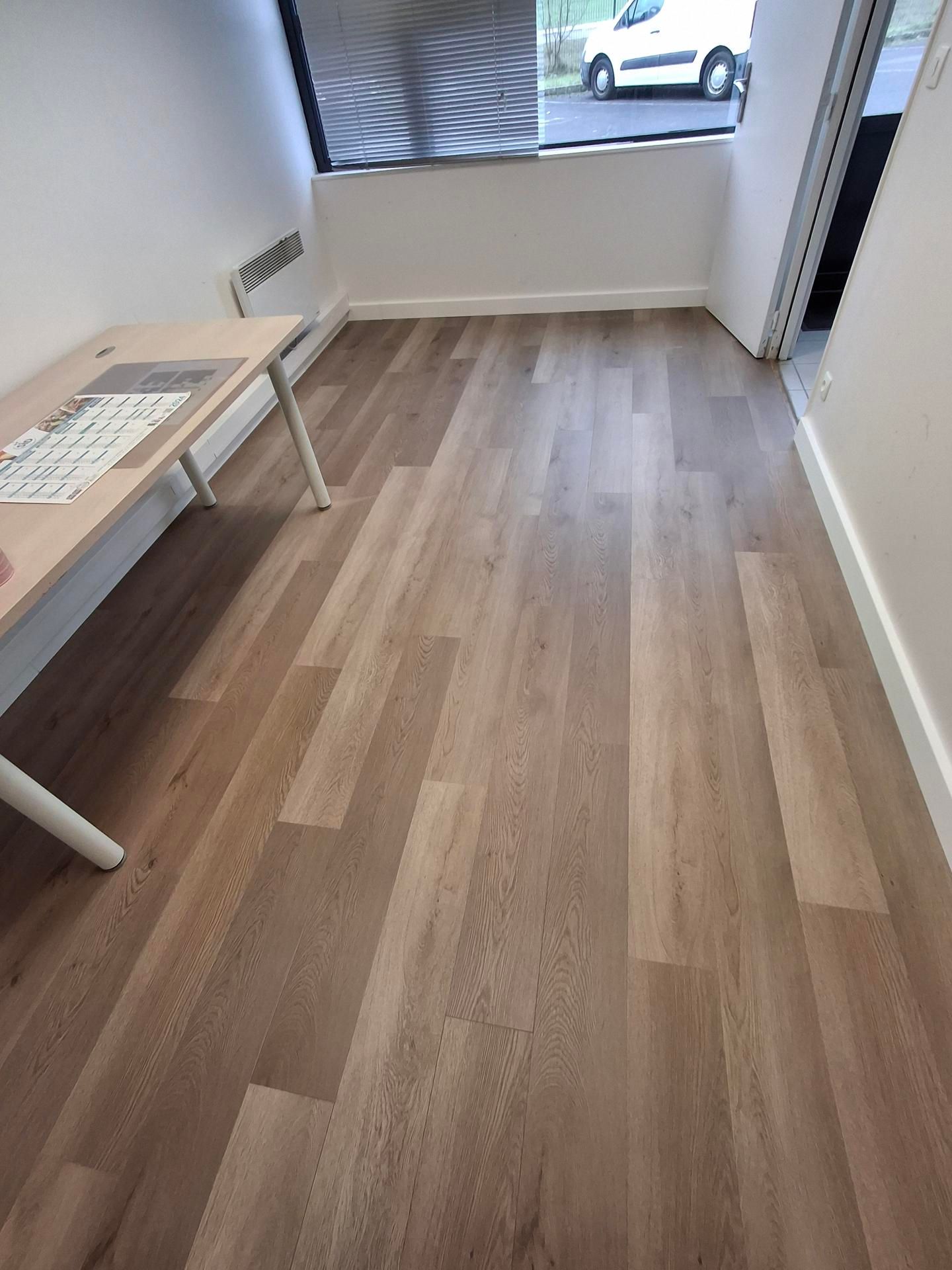 Espace de bureau avec parquet clair et bureau équipé d'un clavier. Fenêtre et porte visibles.