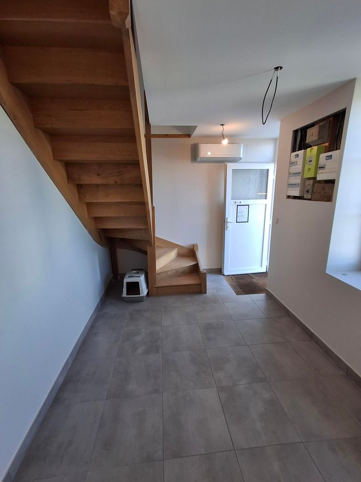 Une entrée lumineuse et étroite avec des escaliers en bois, une porte blanche et un sol carrelé gris.