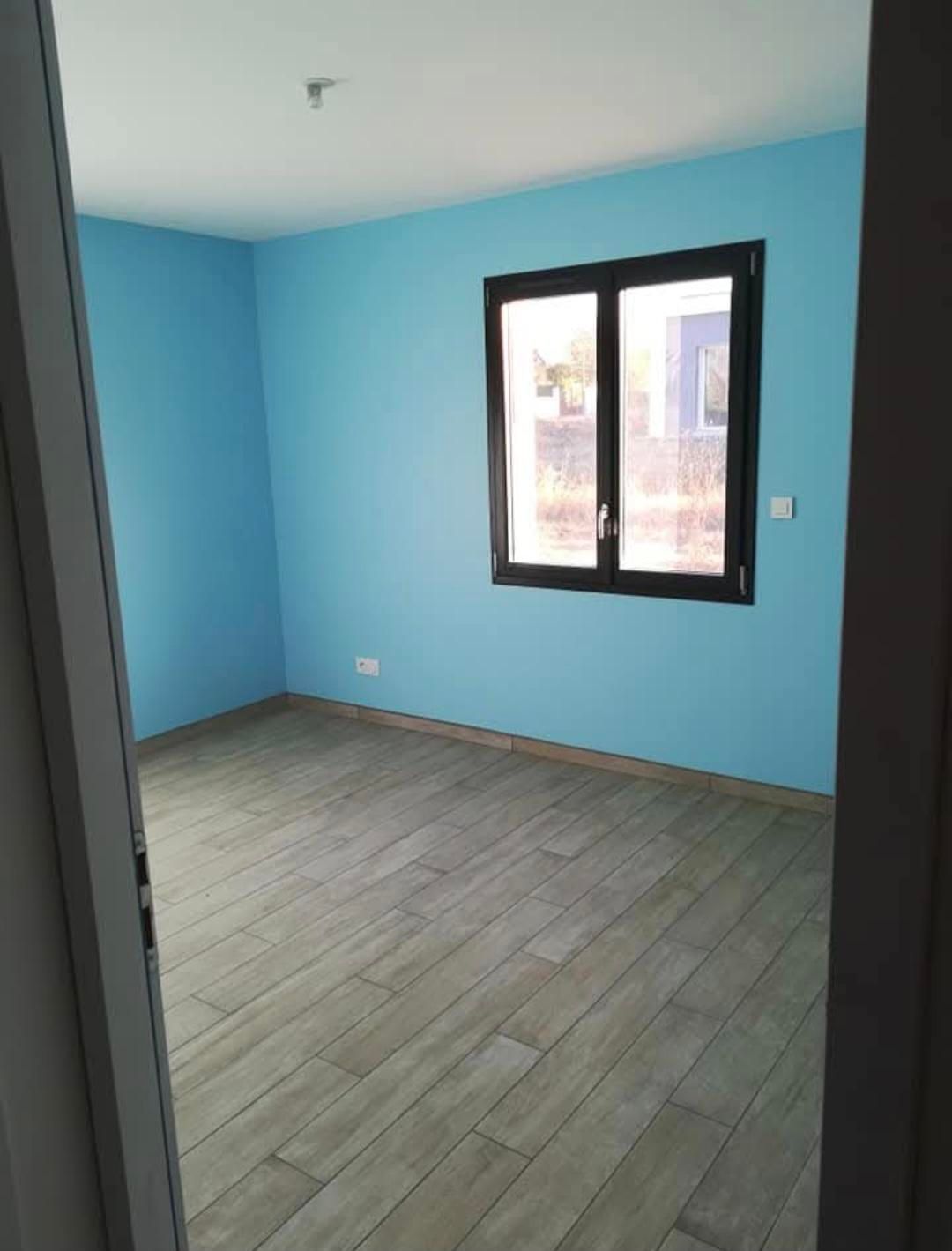 Pièce vide avec des murs bleus, une fenêtre à cadre sombre et un parquet imitation bois.