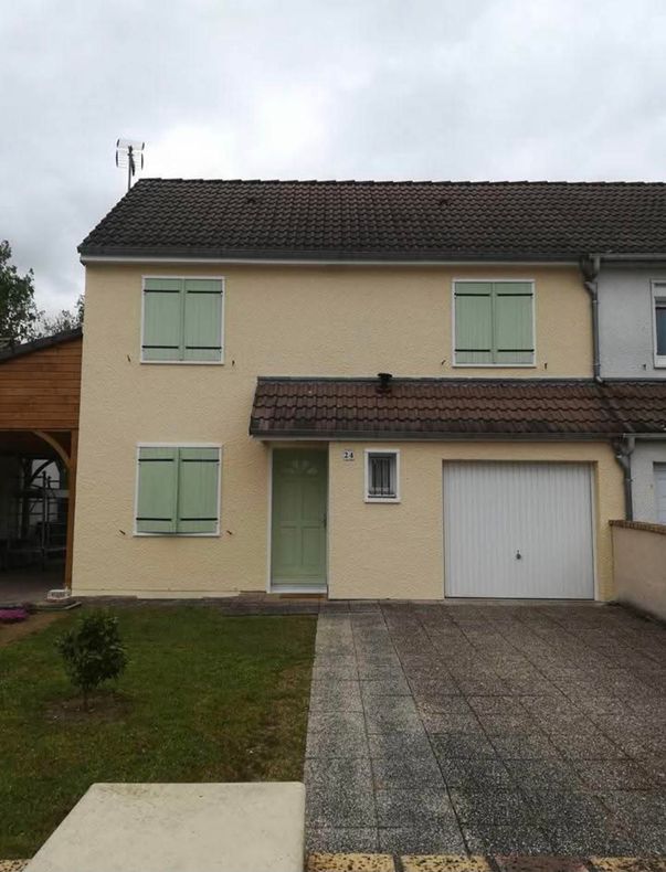 Maison beige à deux étages avec des volets et une porte verts, une porte de garage blanche et une allée grise par temps nuageux.