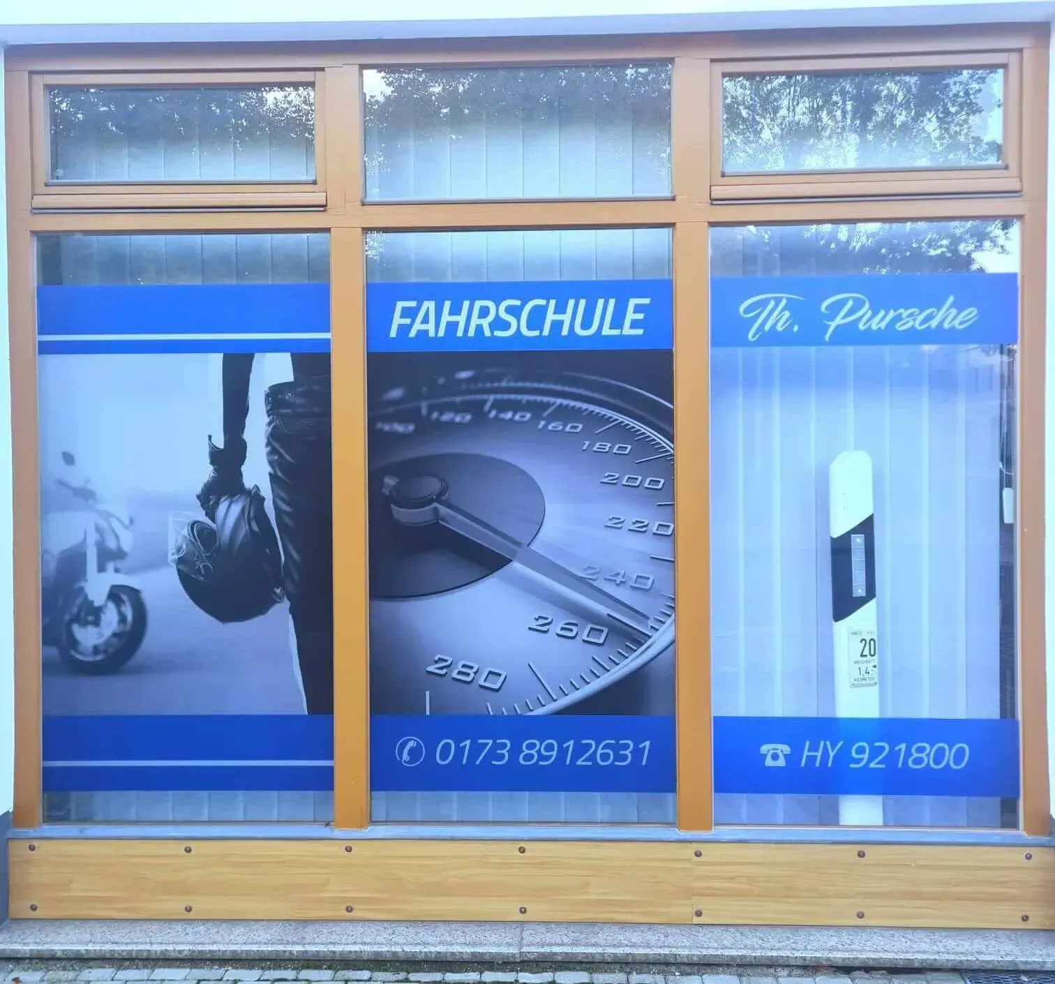 Schaufenster einer Fahrschule mit dem Schriftzug „FAHRSCHULE“ und Kontaktinformationen. Enthält Grafiken eines Motorrads, eines Tachometers und eines Straßenschildes.