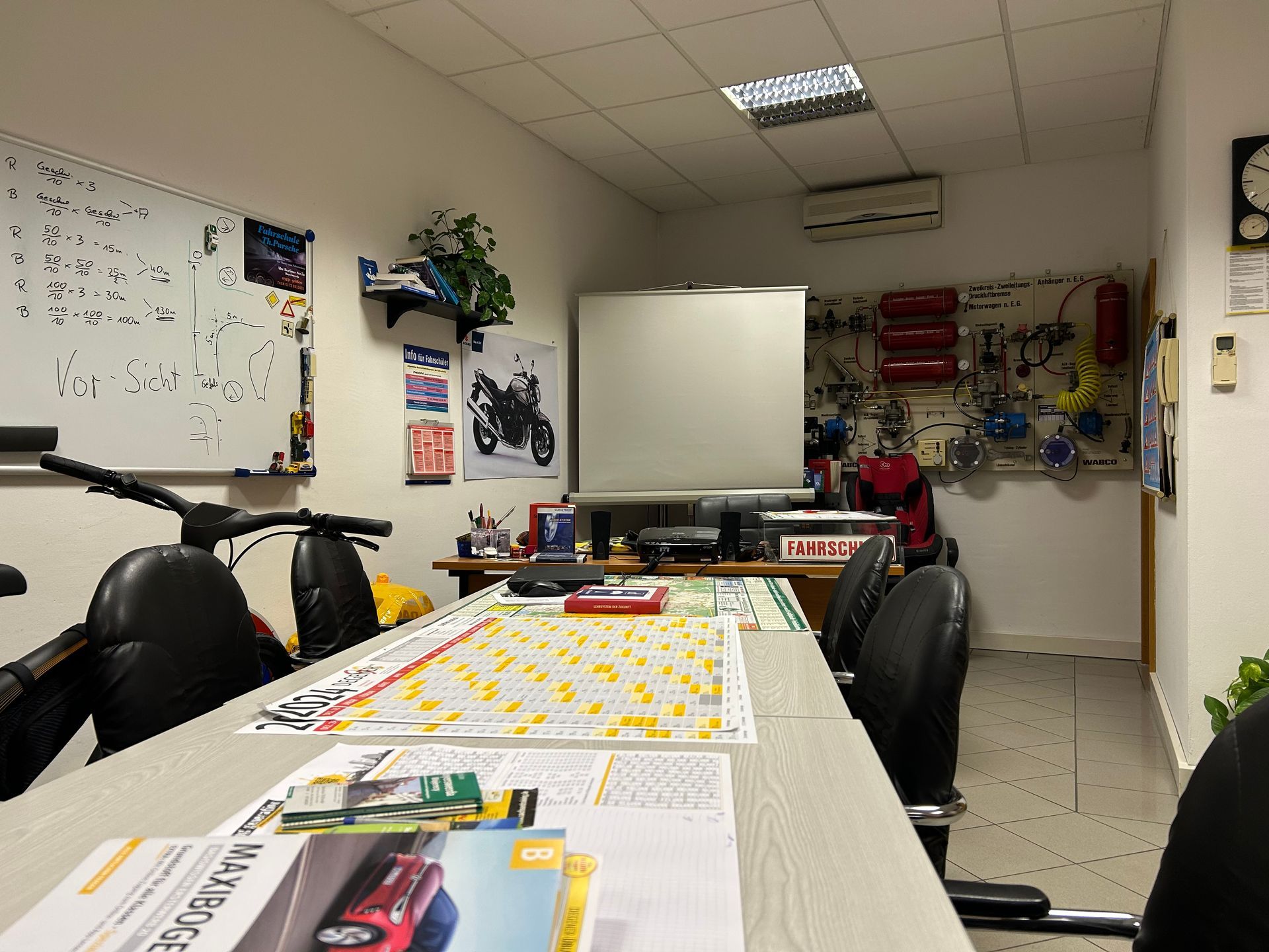 Büro mit Whiteboard, Schreibtisch, Stühlen, Diagrammen und Fahrzeugteilen an den Wänden.