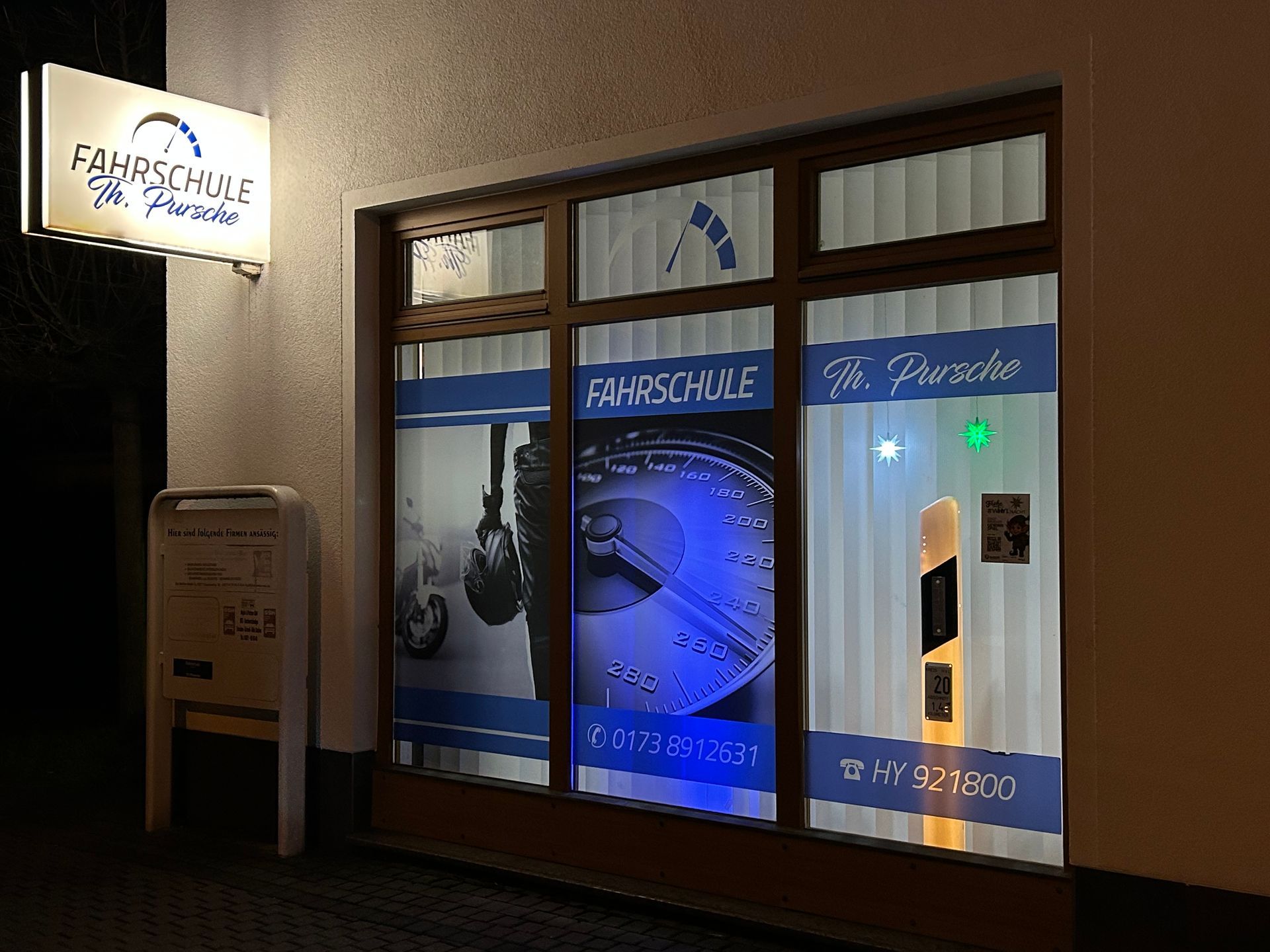 Schaufenster einer Fahrschule bei Nacht mit beleuchtetem Schild und Fensteraufklebern.