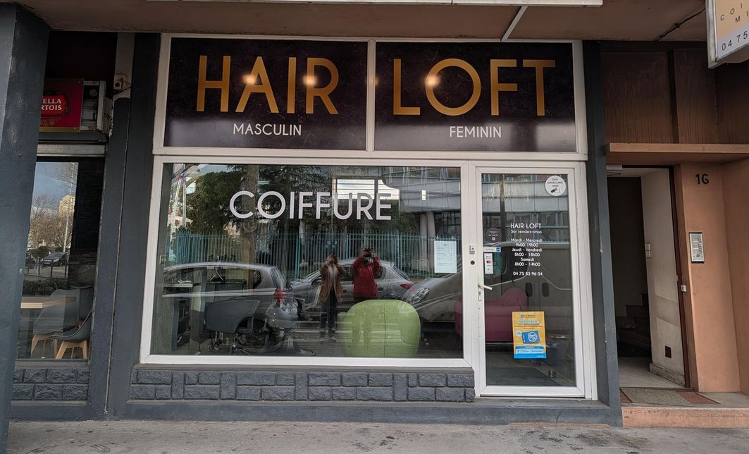 Vue extérieure d'un salon de coiffure nommé HAIR LOFT avec une grande vitrine, une porte d'entrée et un trottoir devant.