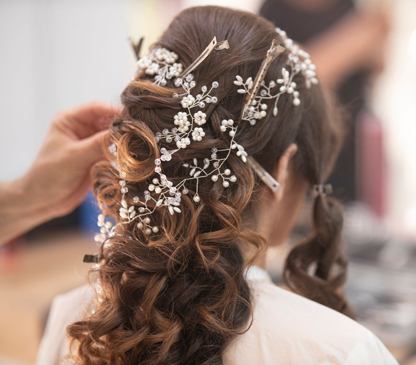 Une coiffeuse arrange des cheveux bruns bouclés ornés d'une délicate parure de tête florale argentée décorée de petites perles blanches.