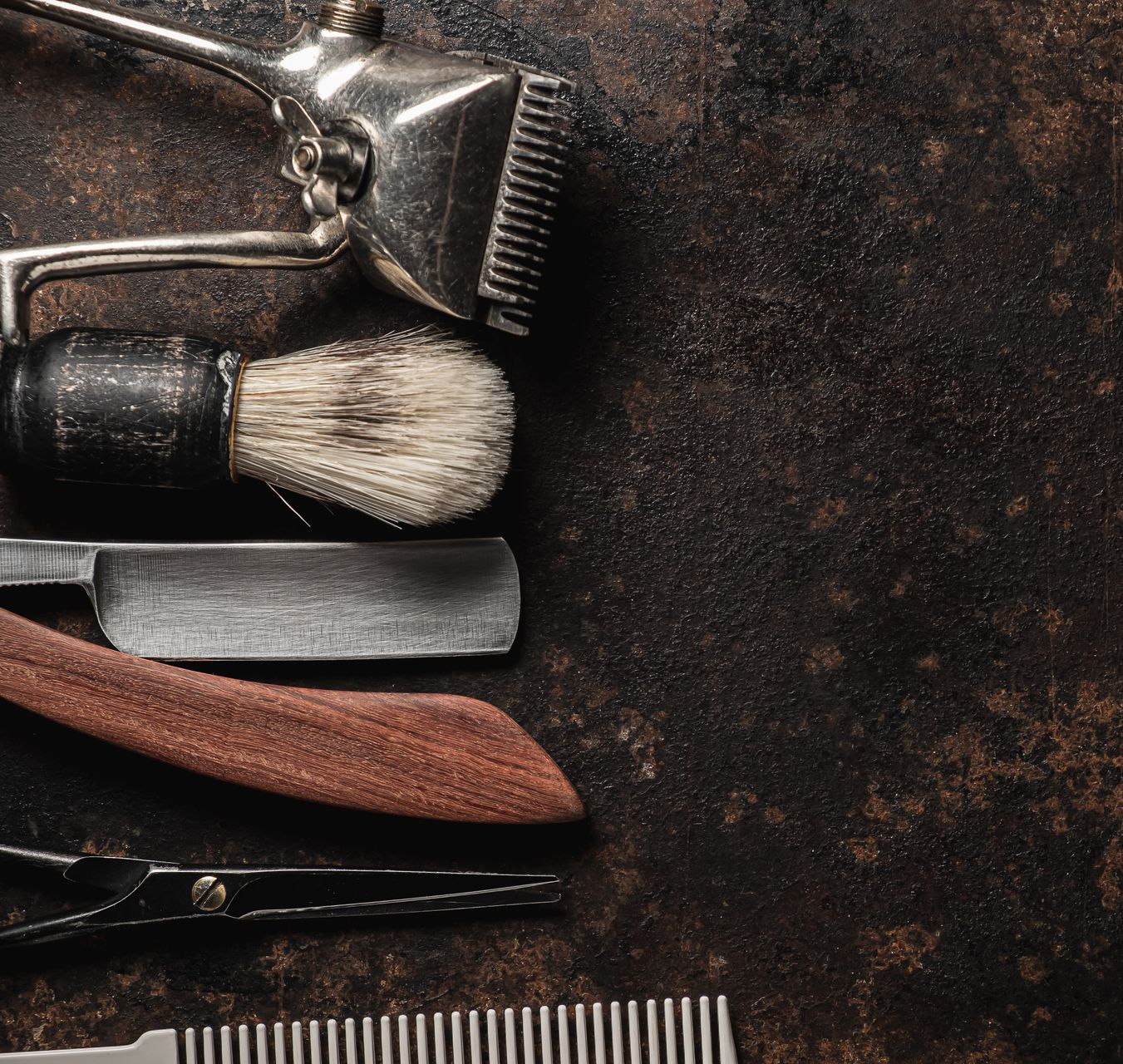 Outils de barbier vintage, notamment une tondeuse, un blaireau, un rasoir droit, des ciseaux et un peigne, sur une surface sombre et rustique.