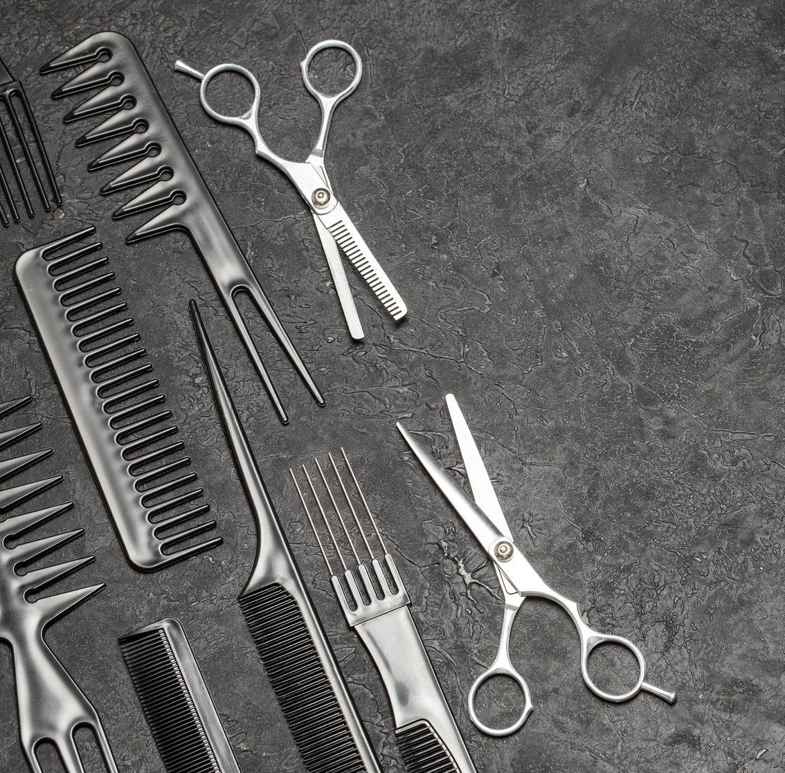 Des outils de coiffure, notamment des peignes et des ciseaux à effiler et à couper, disposés sur une surface sombre et texturée.